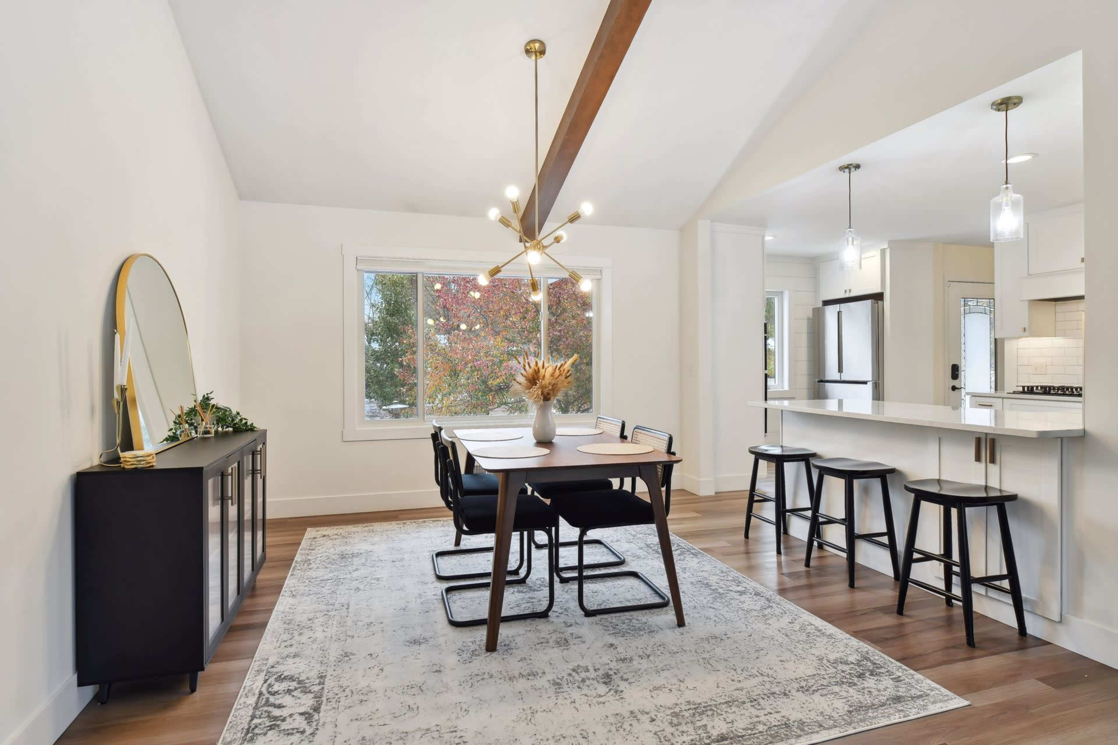 A modern dining area featuring a wooden table with black chairs, a sideboard, and a large window overlooking colorful trees.