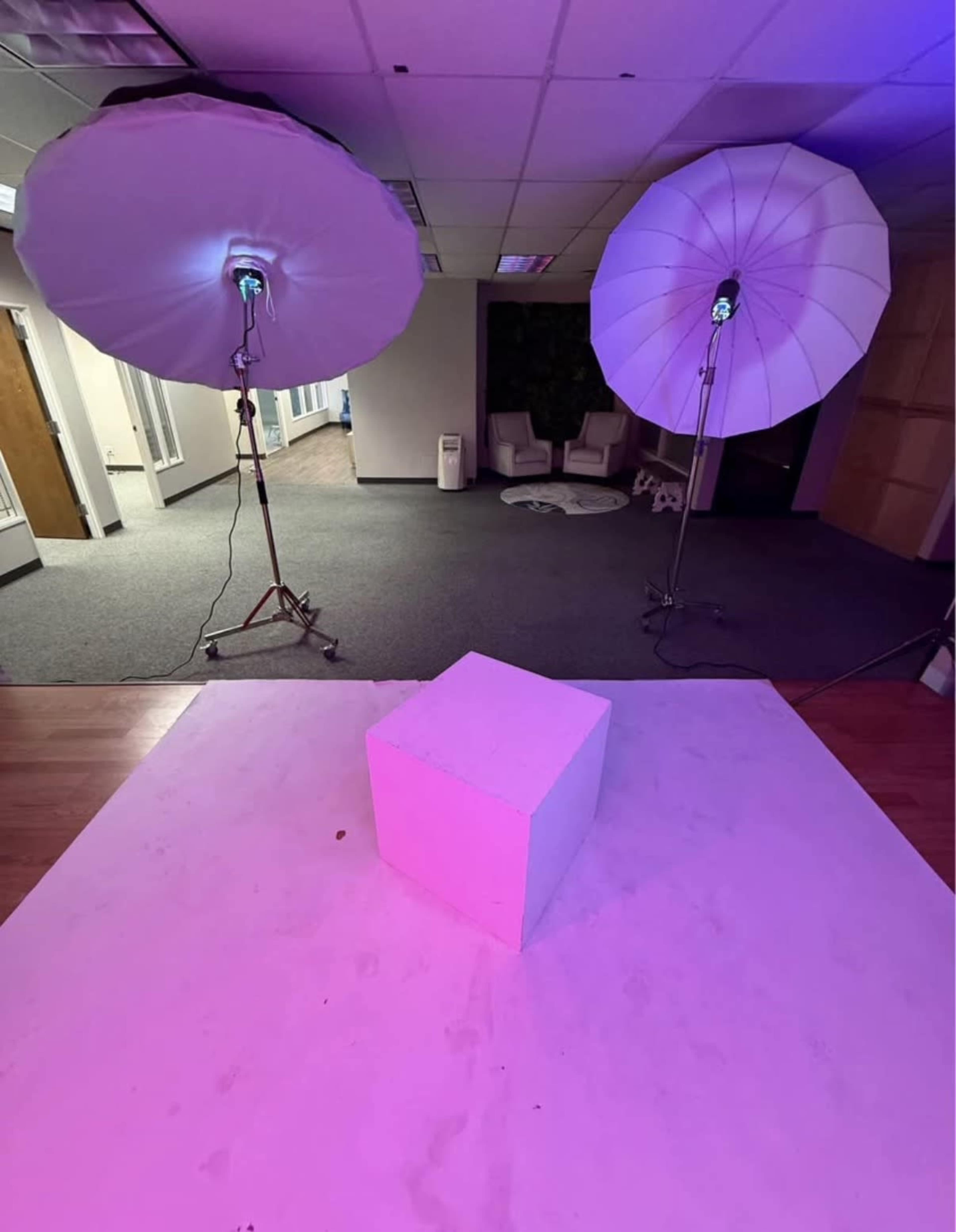 The image shows a minimalistic studio setup with two large softbox lights positioned on either side of a white cube placed on a light-colored surface.