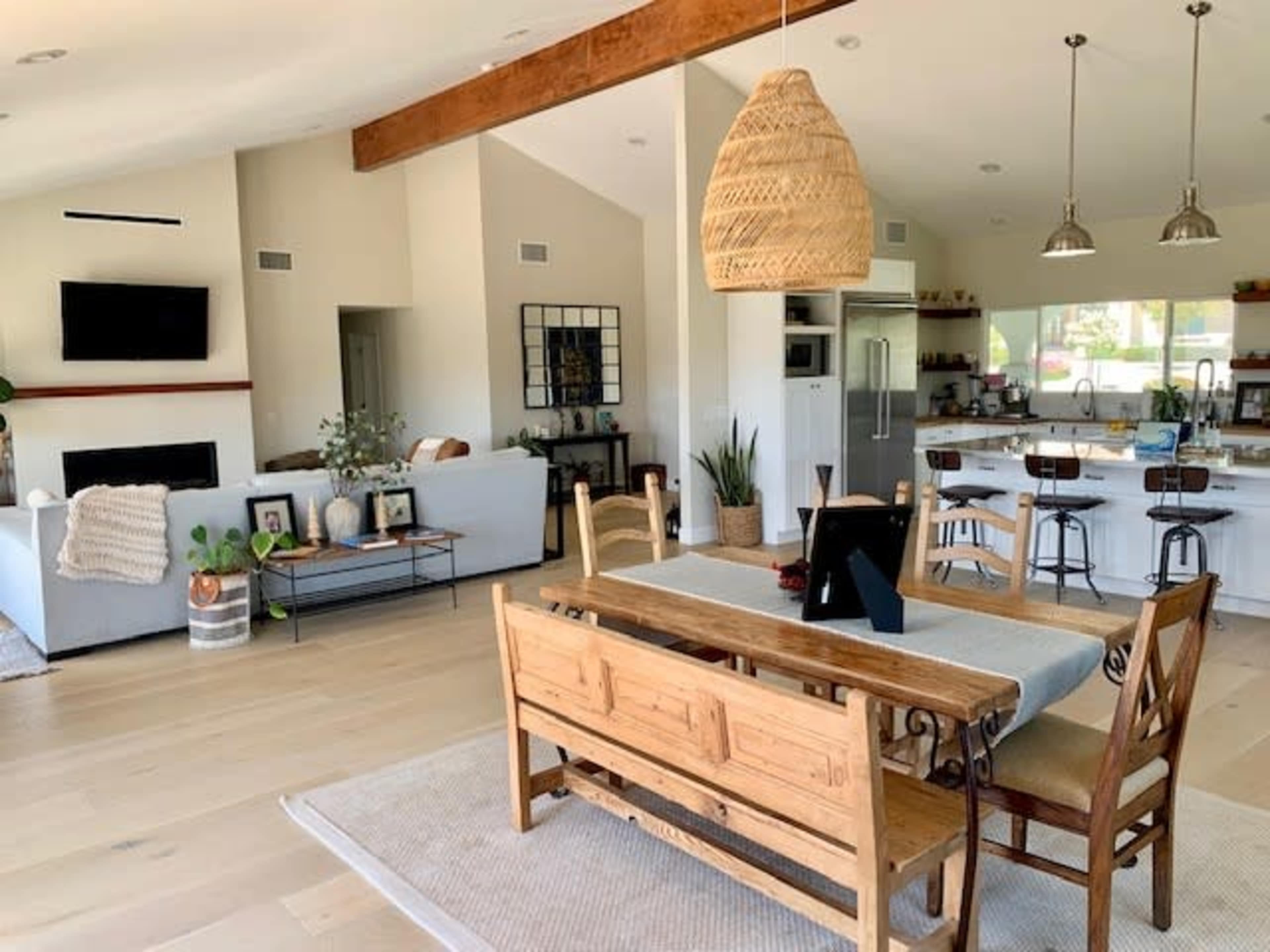A modern interior space featuring a dining area with a wooden table and bench, surrounded by a living room with a sofa and a kitchen in the background.