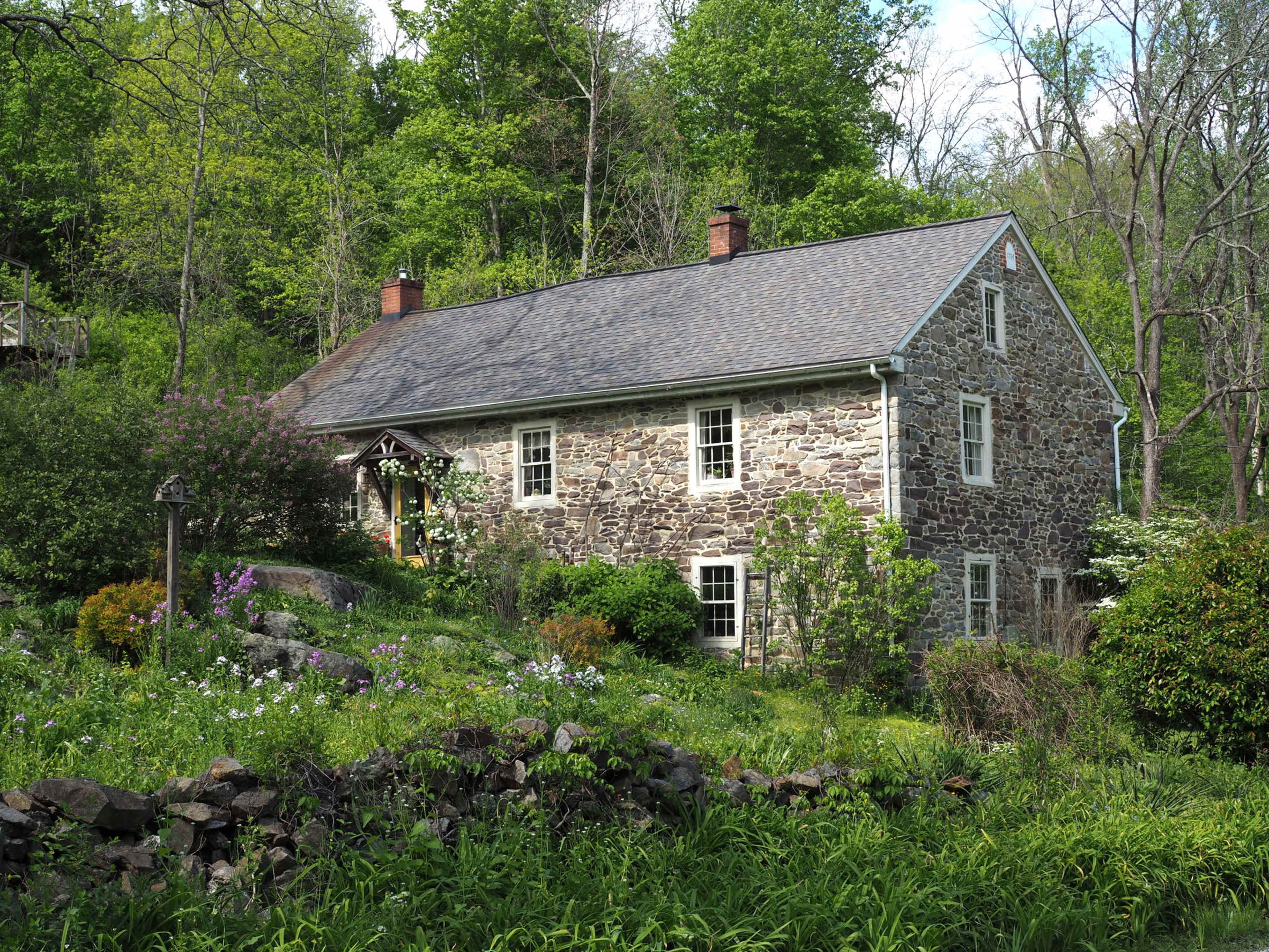 The image shows a stone house surrounded by greenery and flowers in a wooded area.