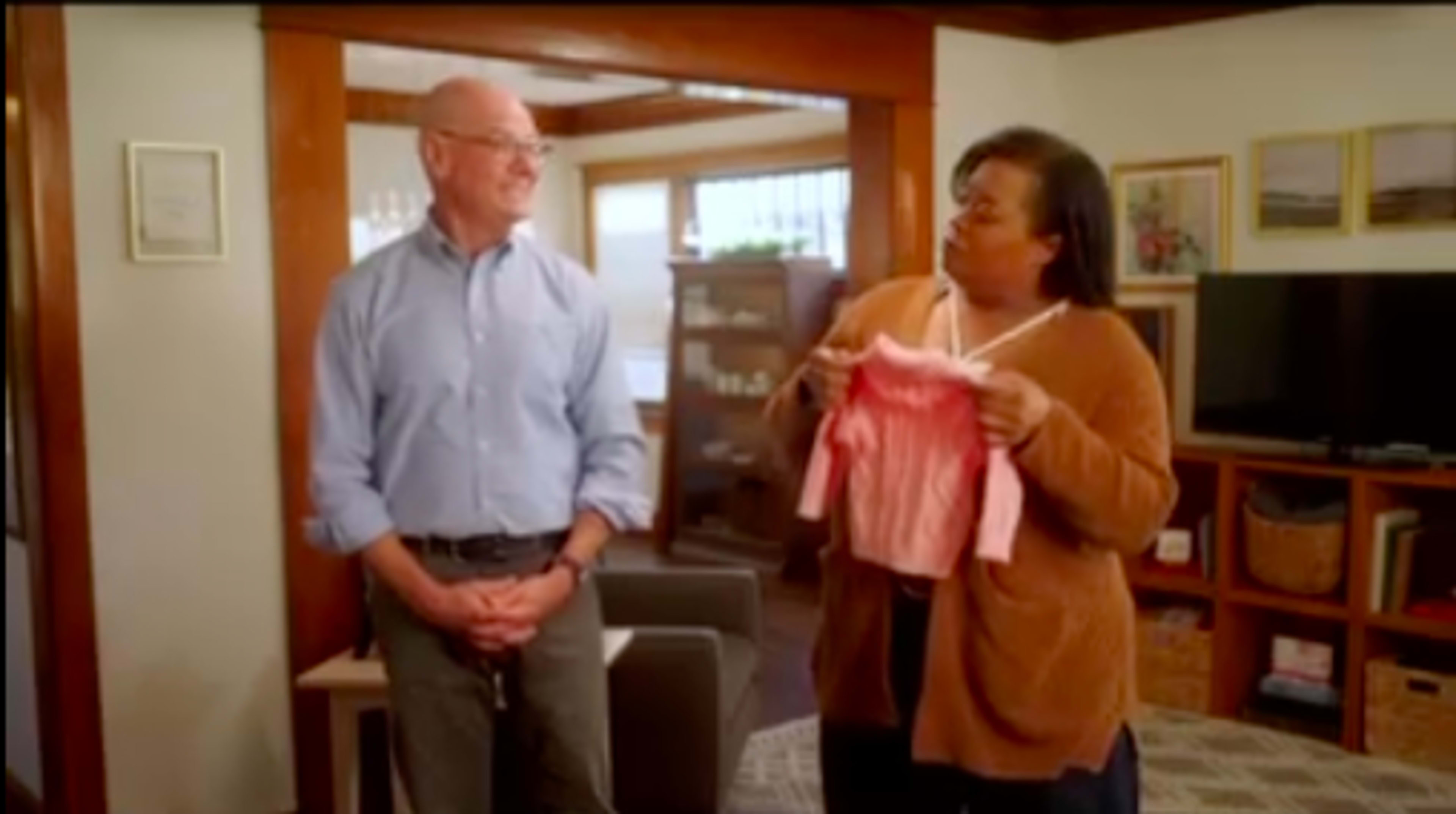 A man stands smiling while a woman holds up a pink garment in a cozy living room setting.