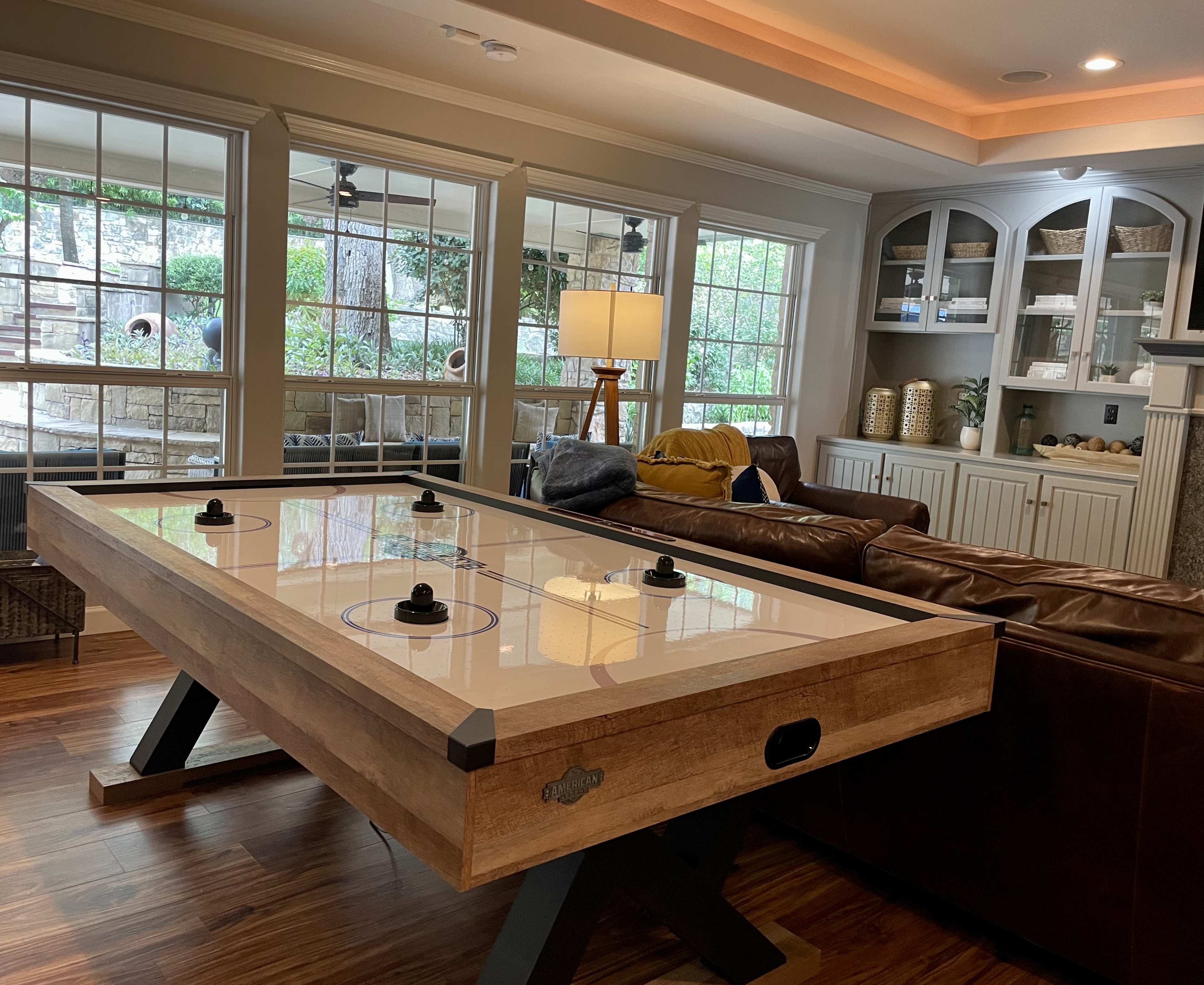 The image shows an air hockey table positioned in a cozy living room with large windows and a sectional sofa.