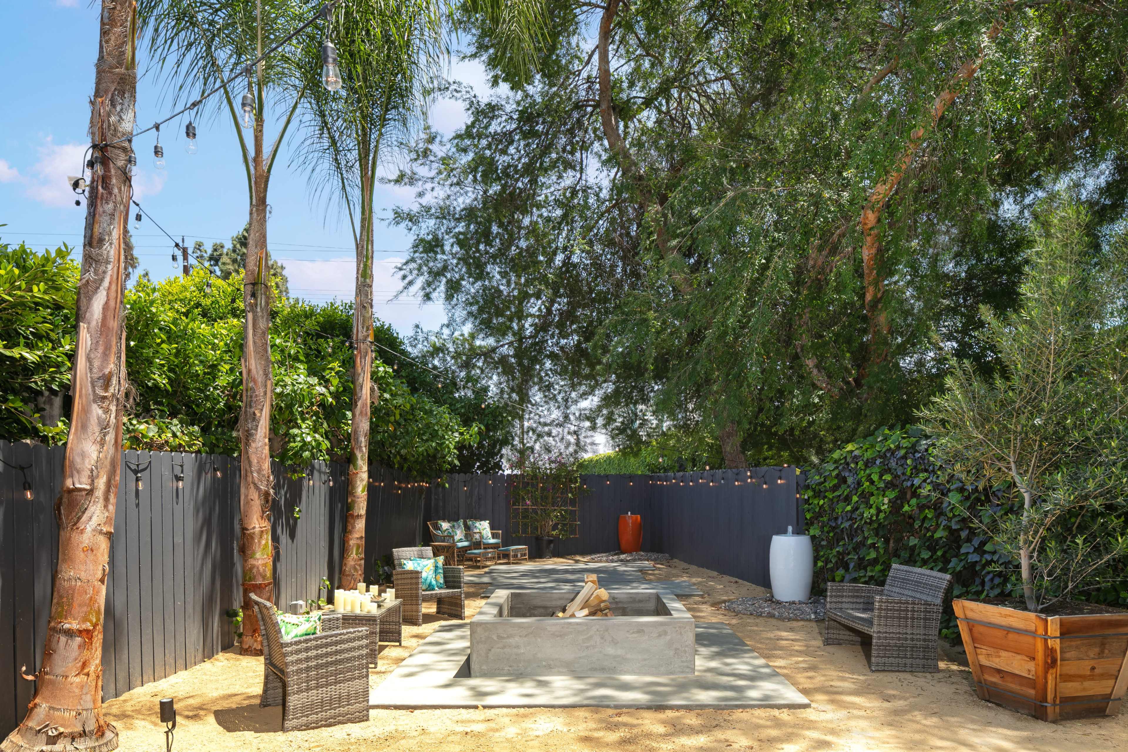 A backyard area with a concrete fire pit surrounded by seating, palm trees, and a dark wooden fence.