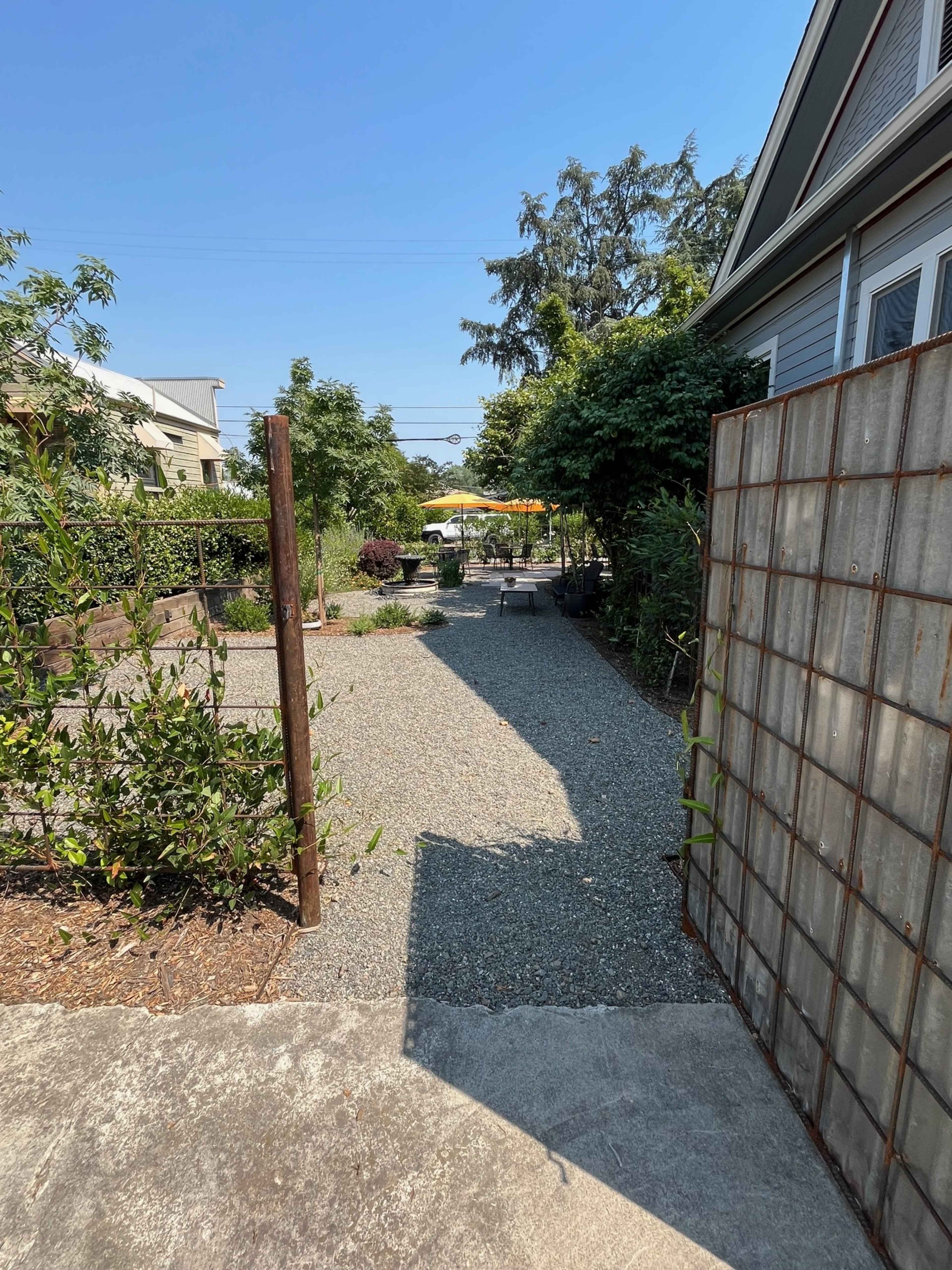 A pathway leads through a gravel yard lined with plants, enclosed by a metal fence, towards a patio area with umbrellas in the distance.