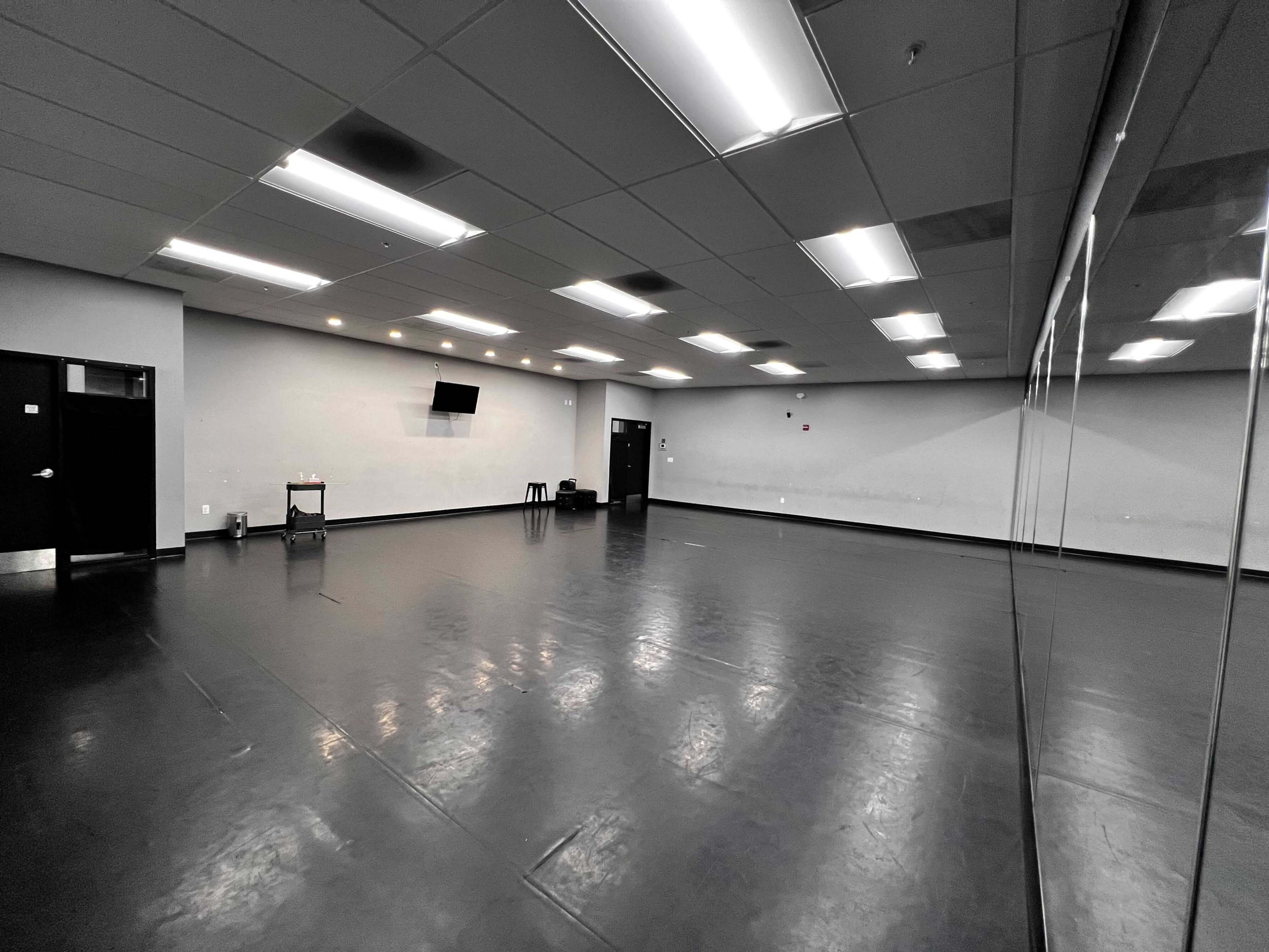 The image shows a spacious, empty room with a reflective wall, overhead lights, and a small table positioned in the corner.