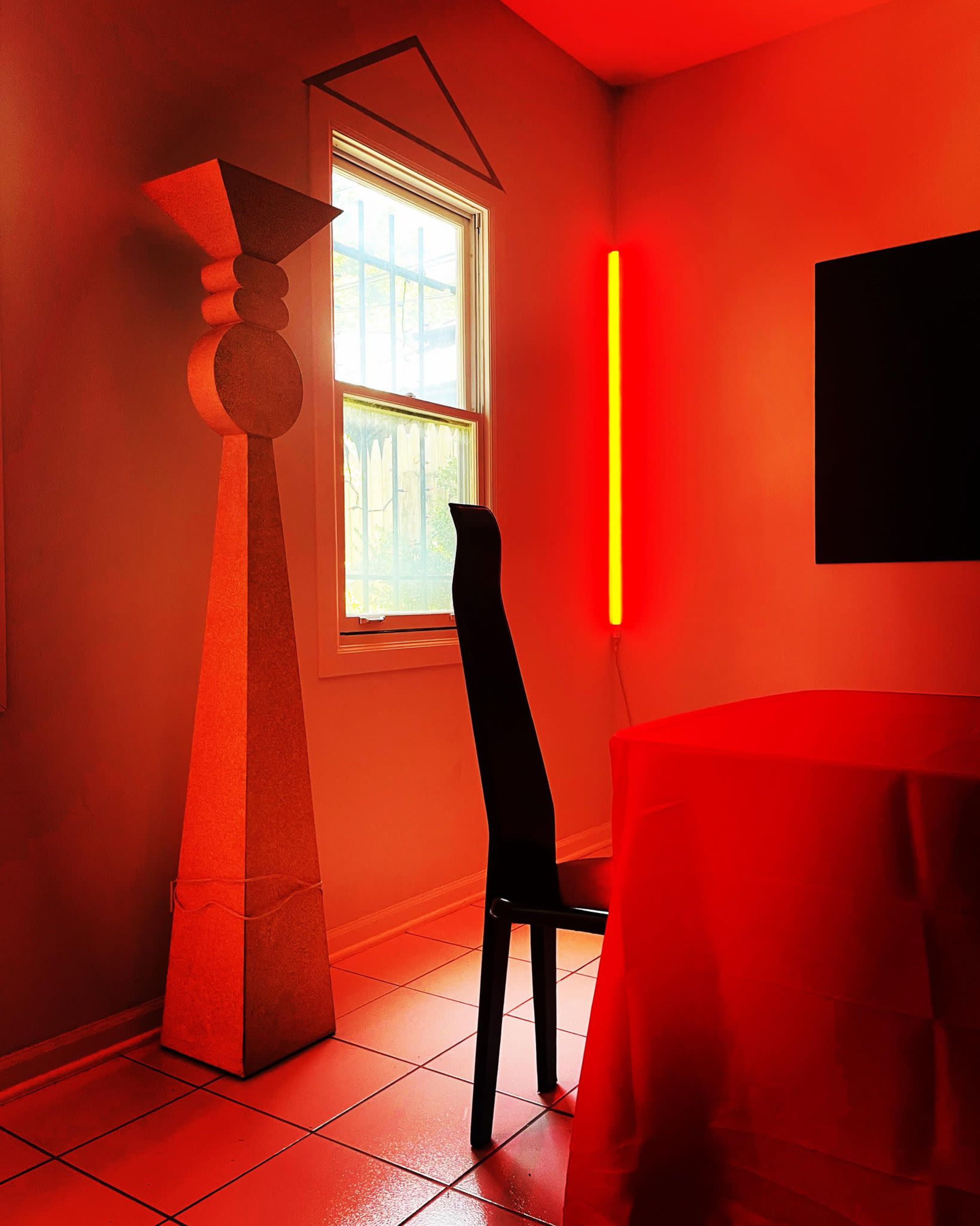 The image shows a room with a red tablecloth, a black chair, a tall sculptural element, and a neon red light illuminating the space near a window.