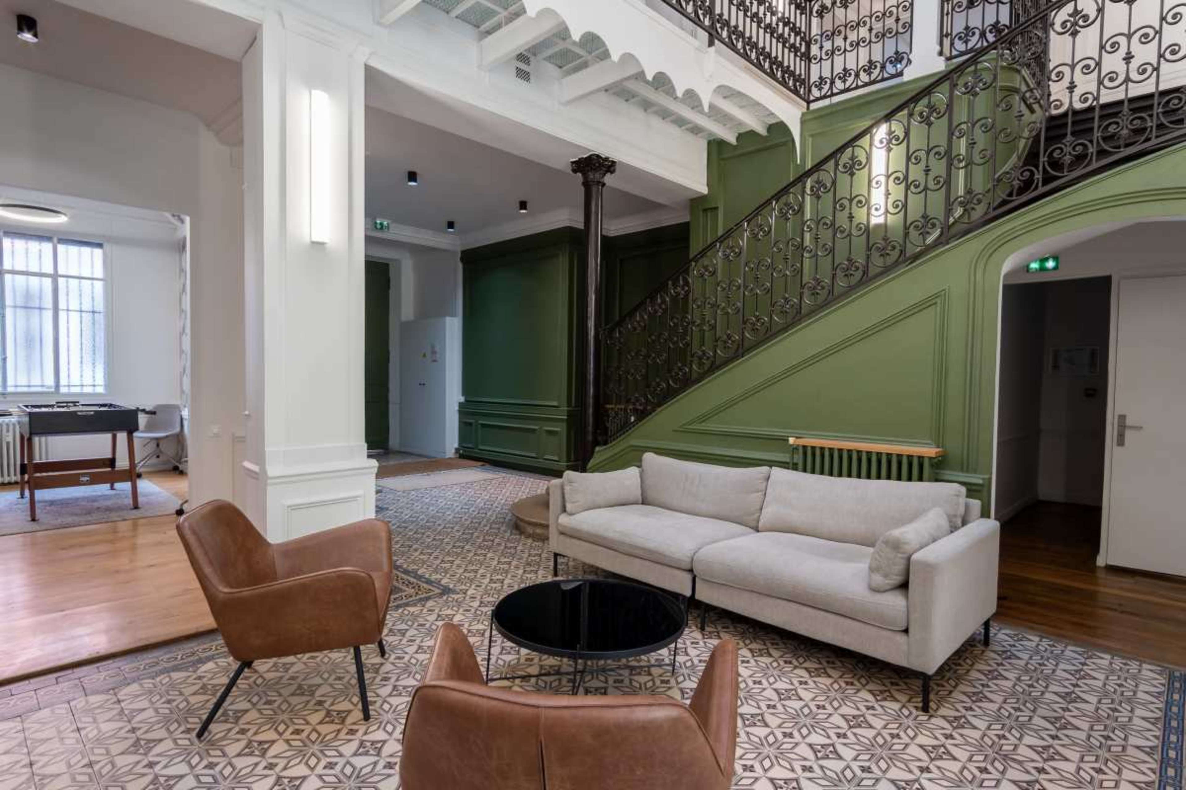 The image shows a well-lit lobby area with a staircase, a gray sofa, two brown chairs, and patterned tile flooring.
