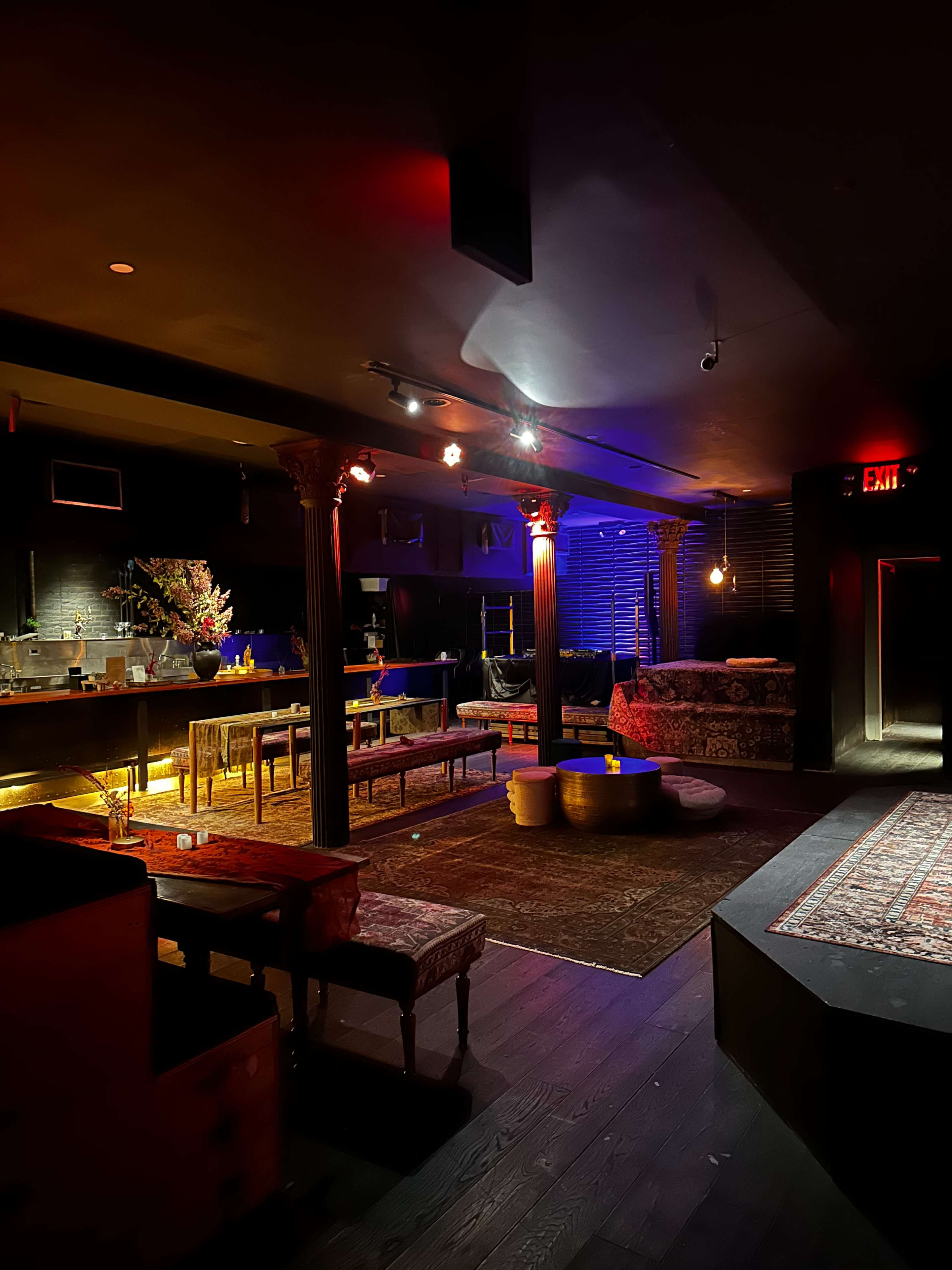 The image shows a dimly lit lounge with elegant seating, decorative pillars, and an illuminated bar area.