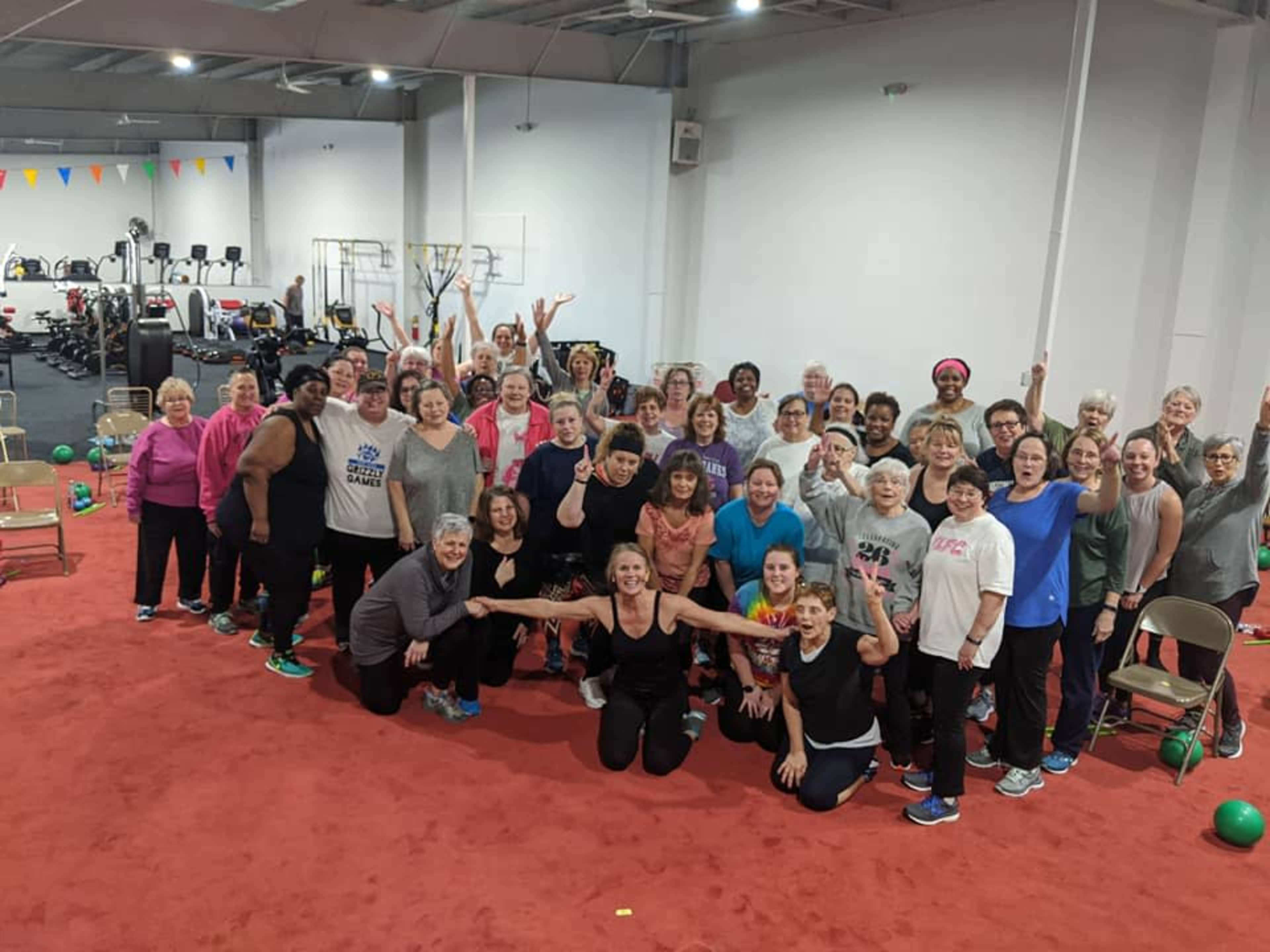 A large group of women of various ages poses together in a fitness center, with exercise equipment visible in the background.