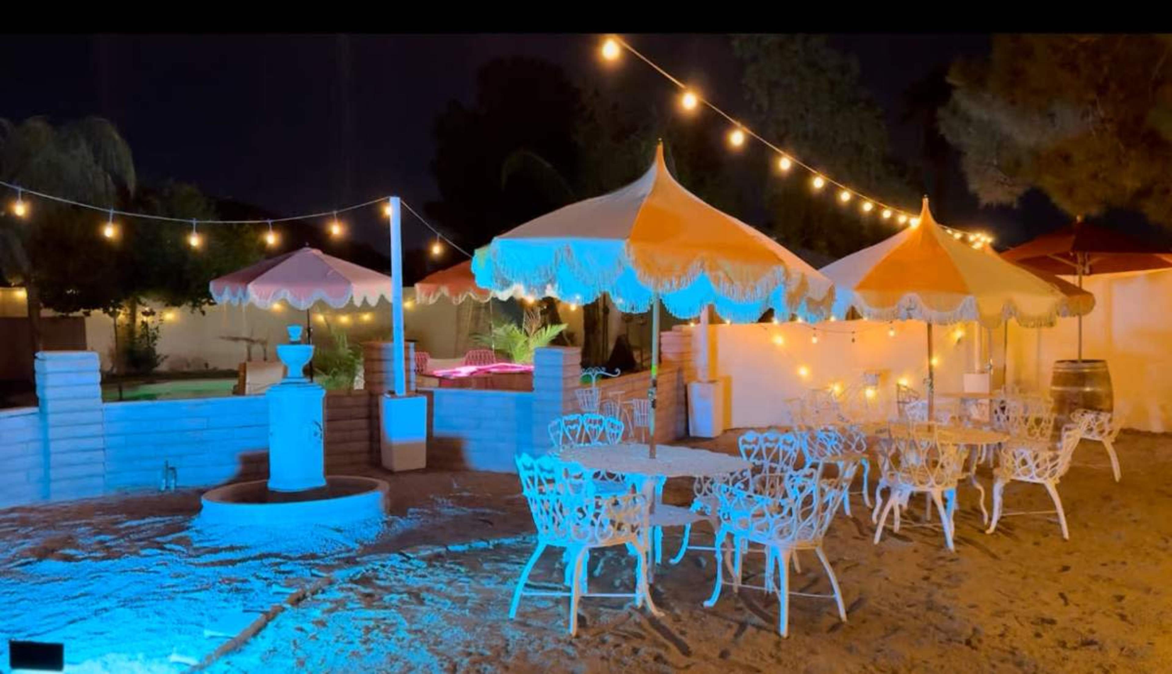 The image shows a nighttime outdoor setting with string lights illuminating white metal tables and chairs under colorful umbrellas near a pool.