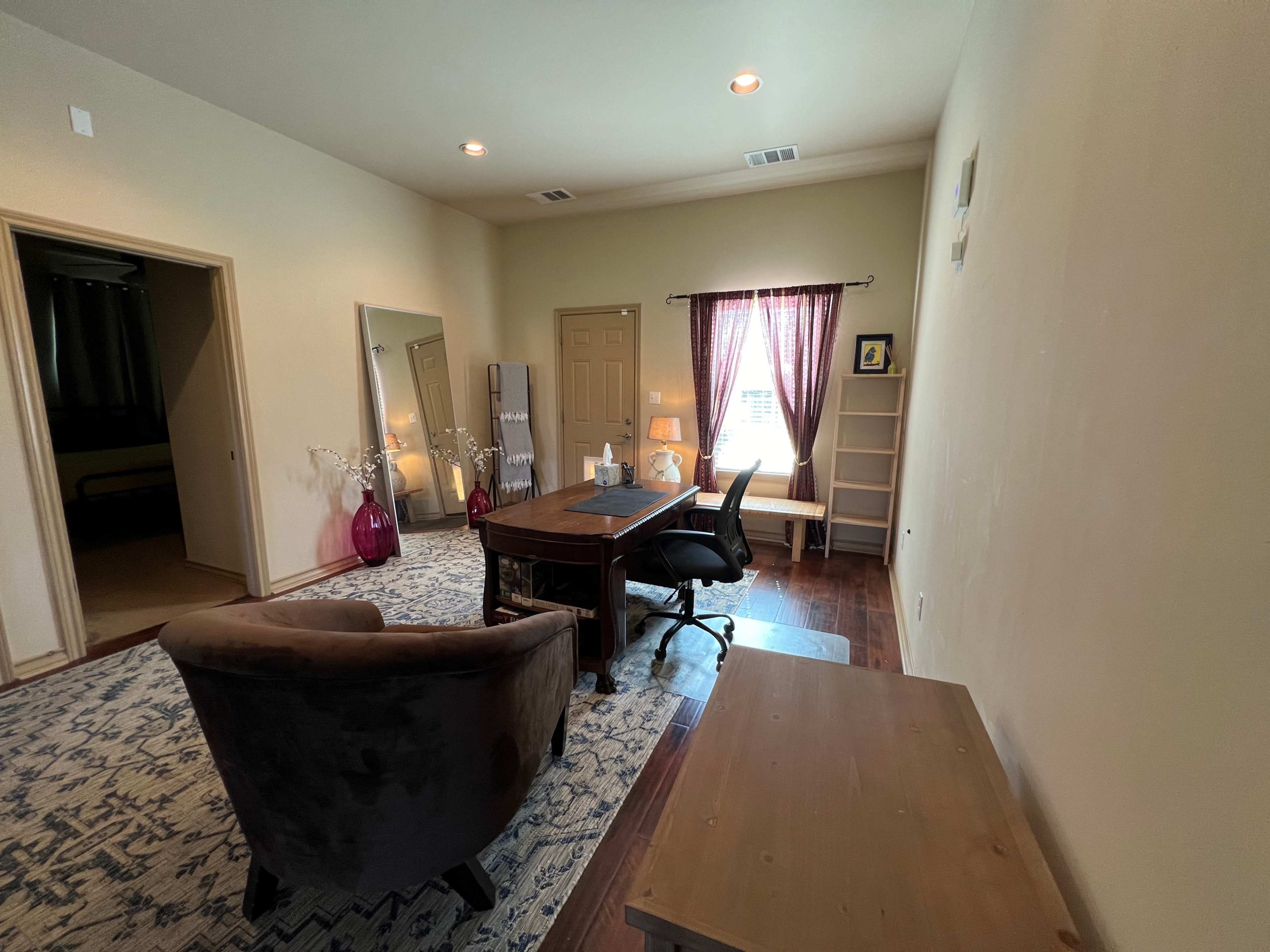 A spacious office featuring a desk and chair, a mirror, a bookshelf, and decorative elements, with natural light filtering through a curtain.