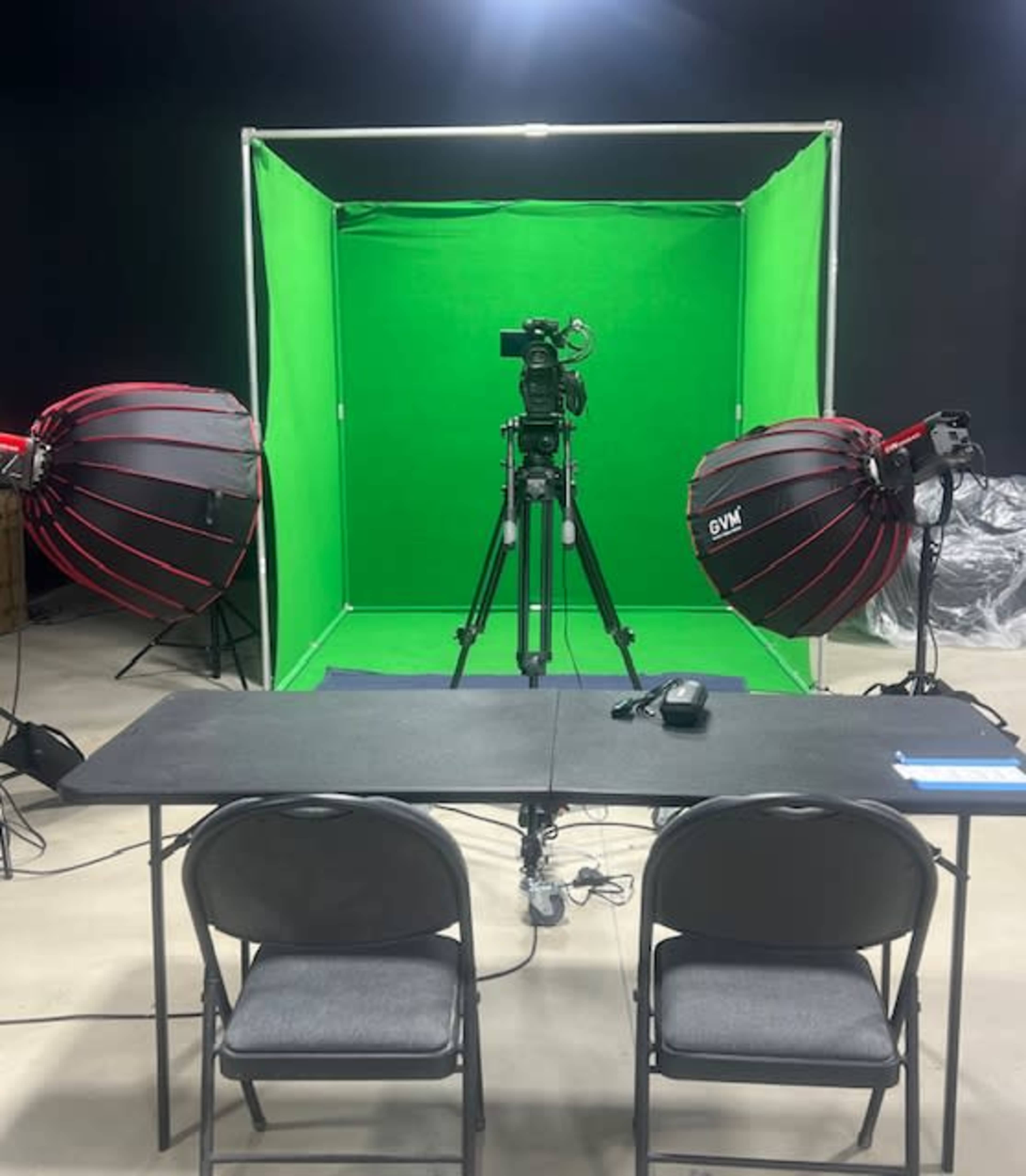 A green screen setup featuring a camera on a tripod and two large light sources positioned on either side of a black table and chairs.