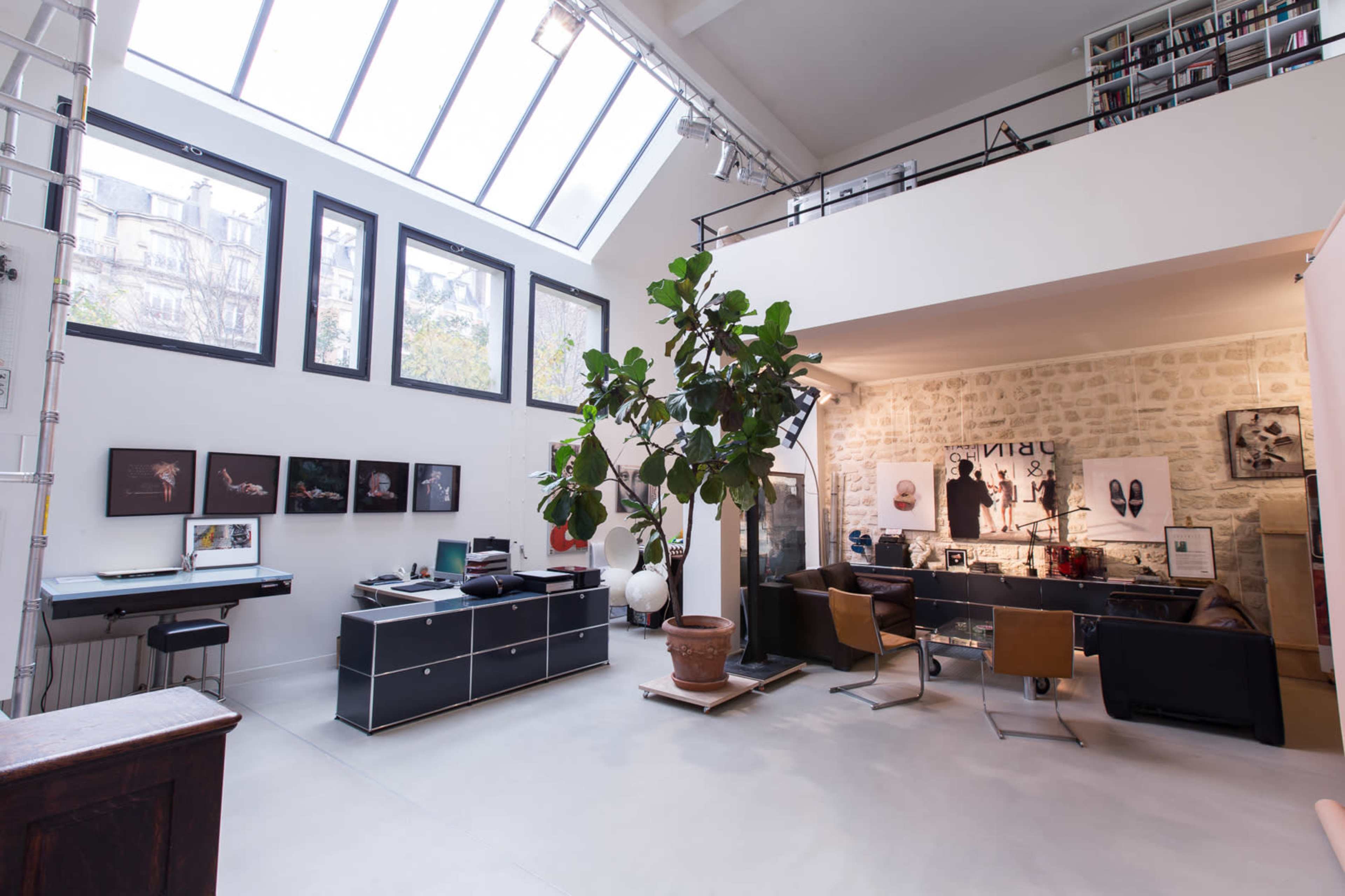 A modern open-concept office space featuring large windows, a central potted tree, and various desks with artwork displayed on the walls.