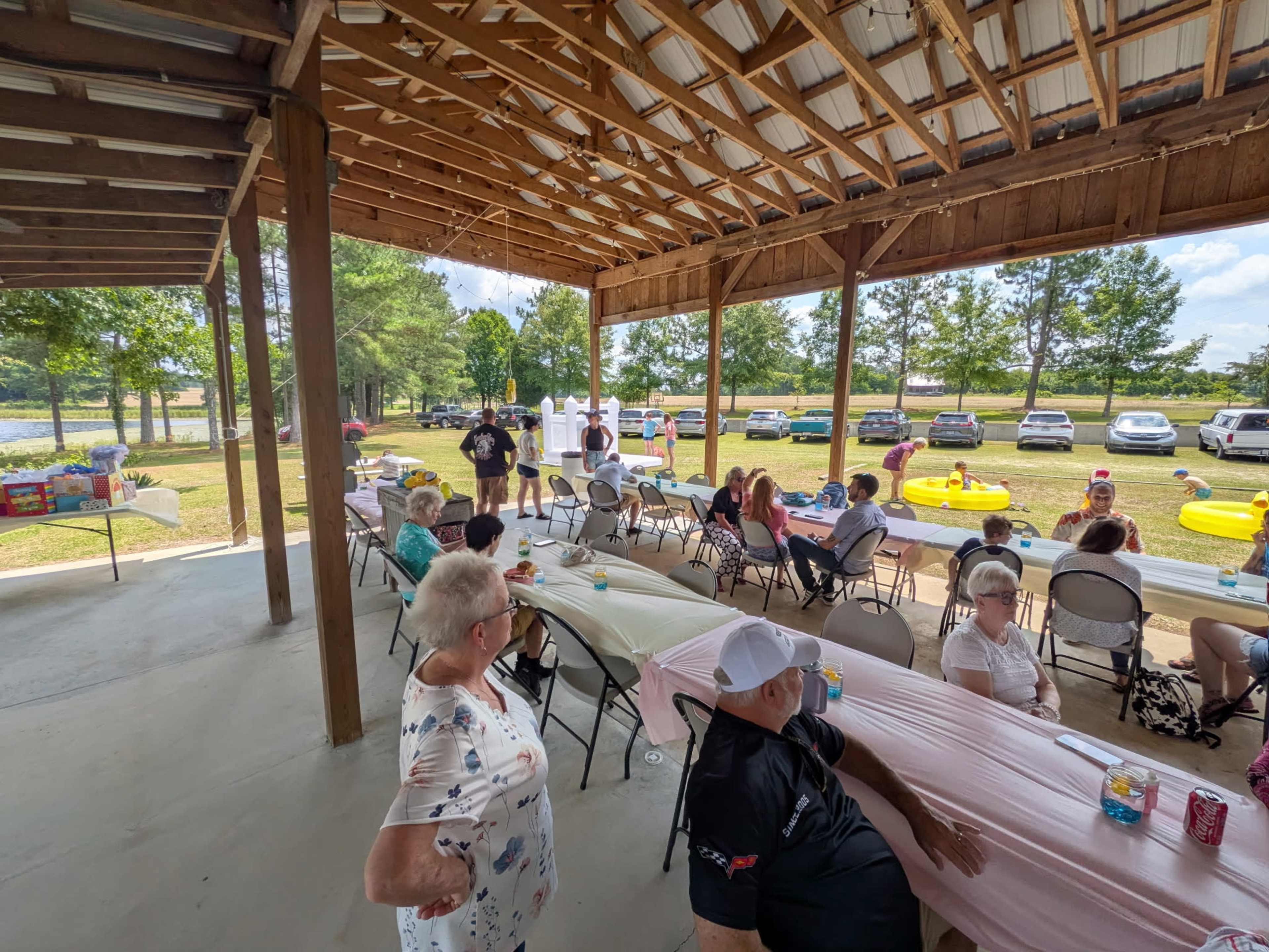 The image shows a gathering under a covered outdoor area with several tables and chairs set up, where people are socializing, while cars and inflatable pool toys are visible in the background.