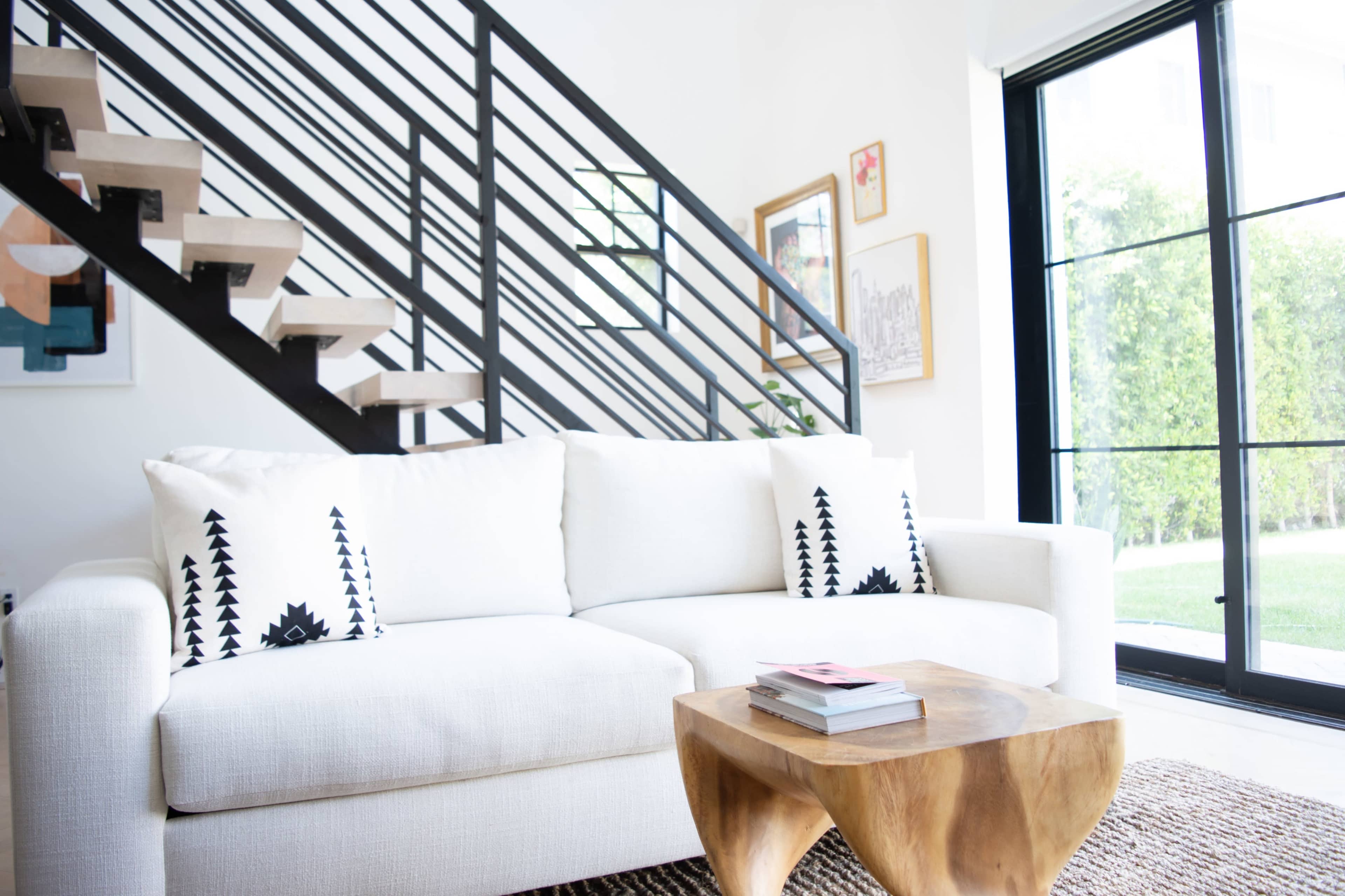 A white sofa with patterned cushions is positioned in a bright room beside a wooden coffee table and a staircase with black railings.