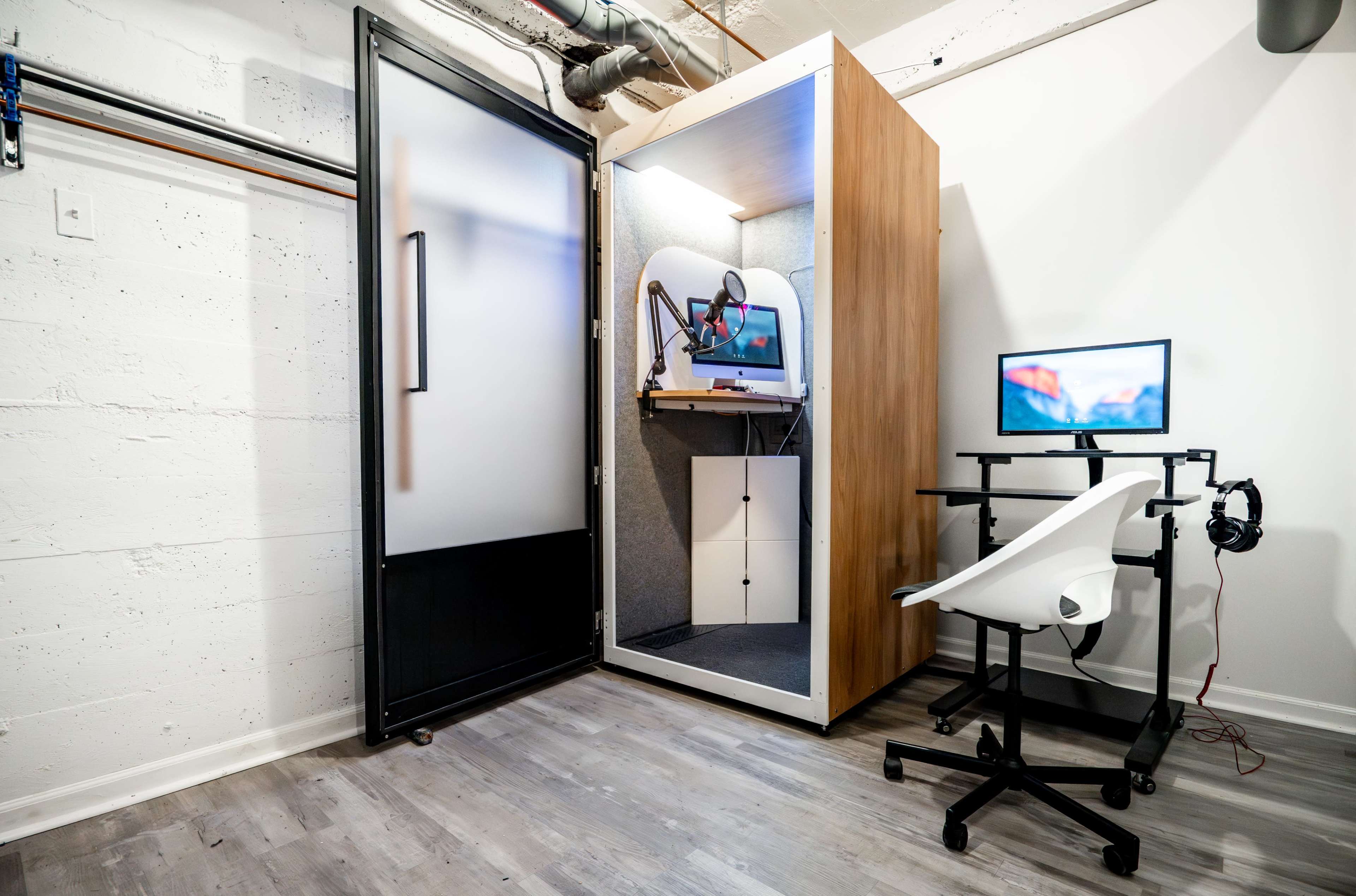 The image shows a modern workspace featuring a soundproof booth with a computer setup inside it, alongside a desk with a monitor and headset.