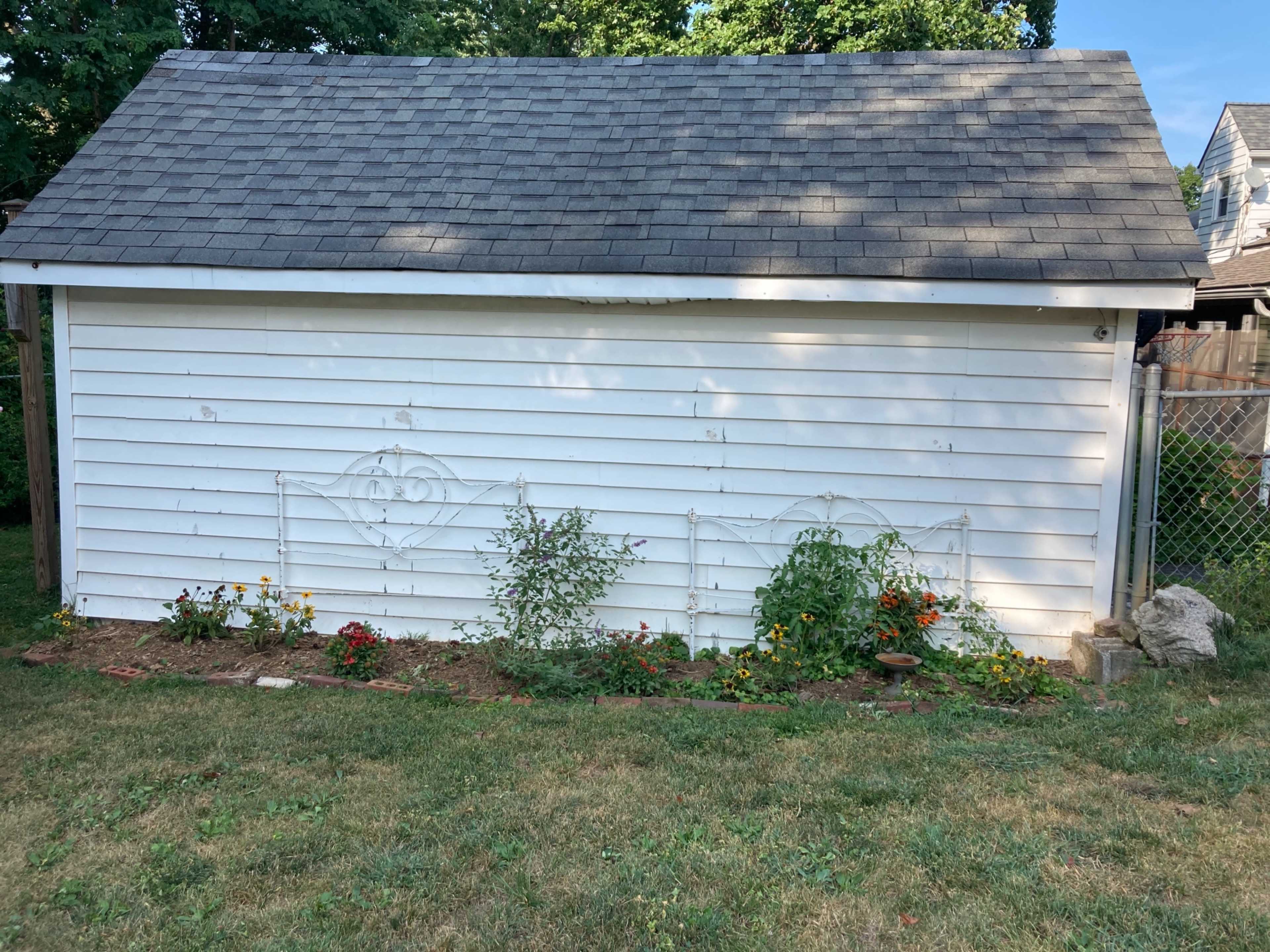 A white shed with a gray roof has a flowerbed lined with various plants and flowers in front of it.