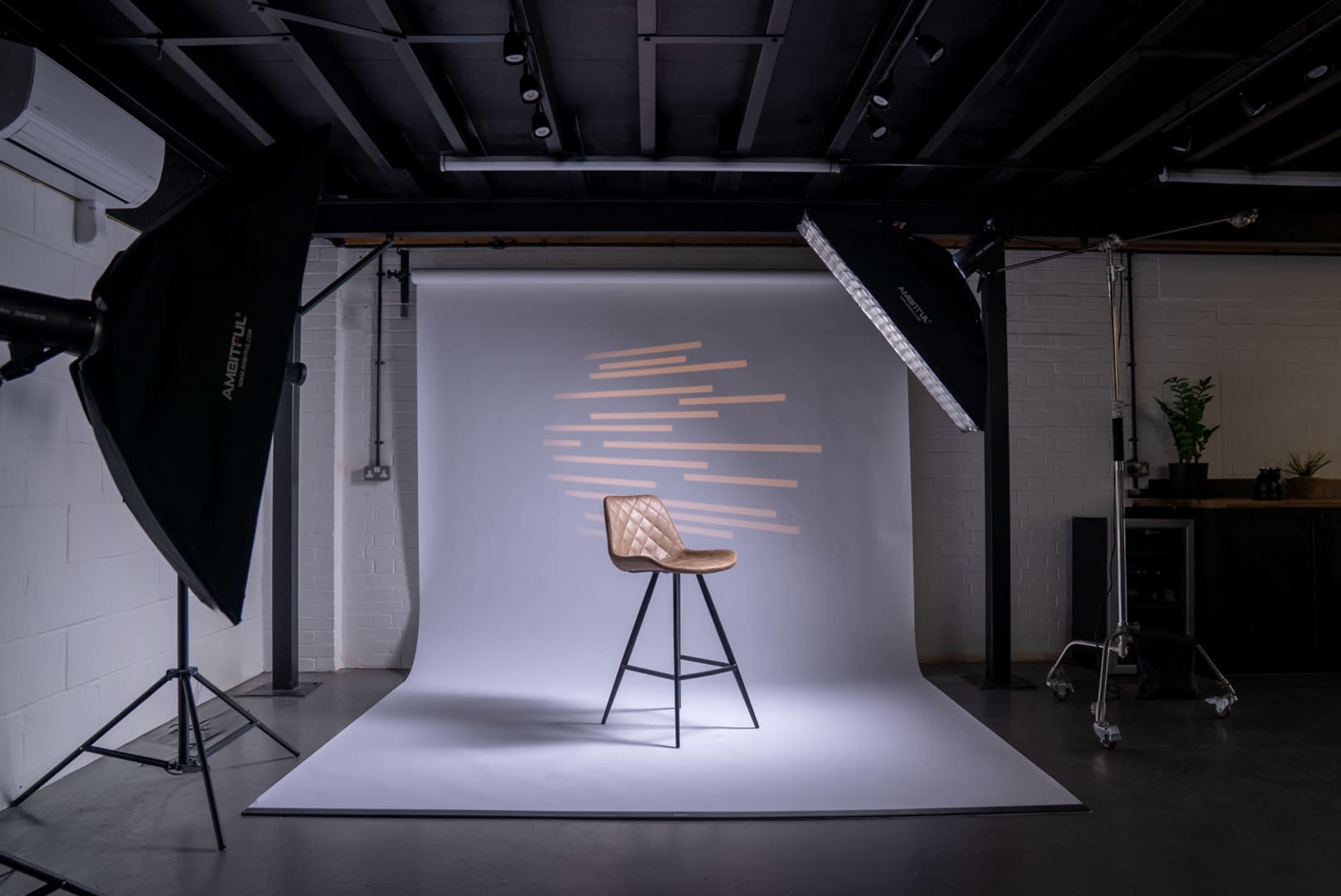 The image shows a photography studio featuring a single wooden stool against a smooth backdrop with soft lighting and geometric shadow patterns.