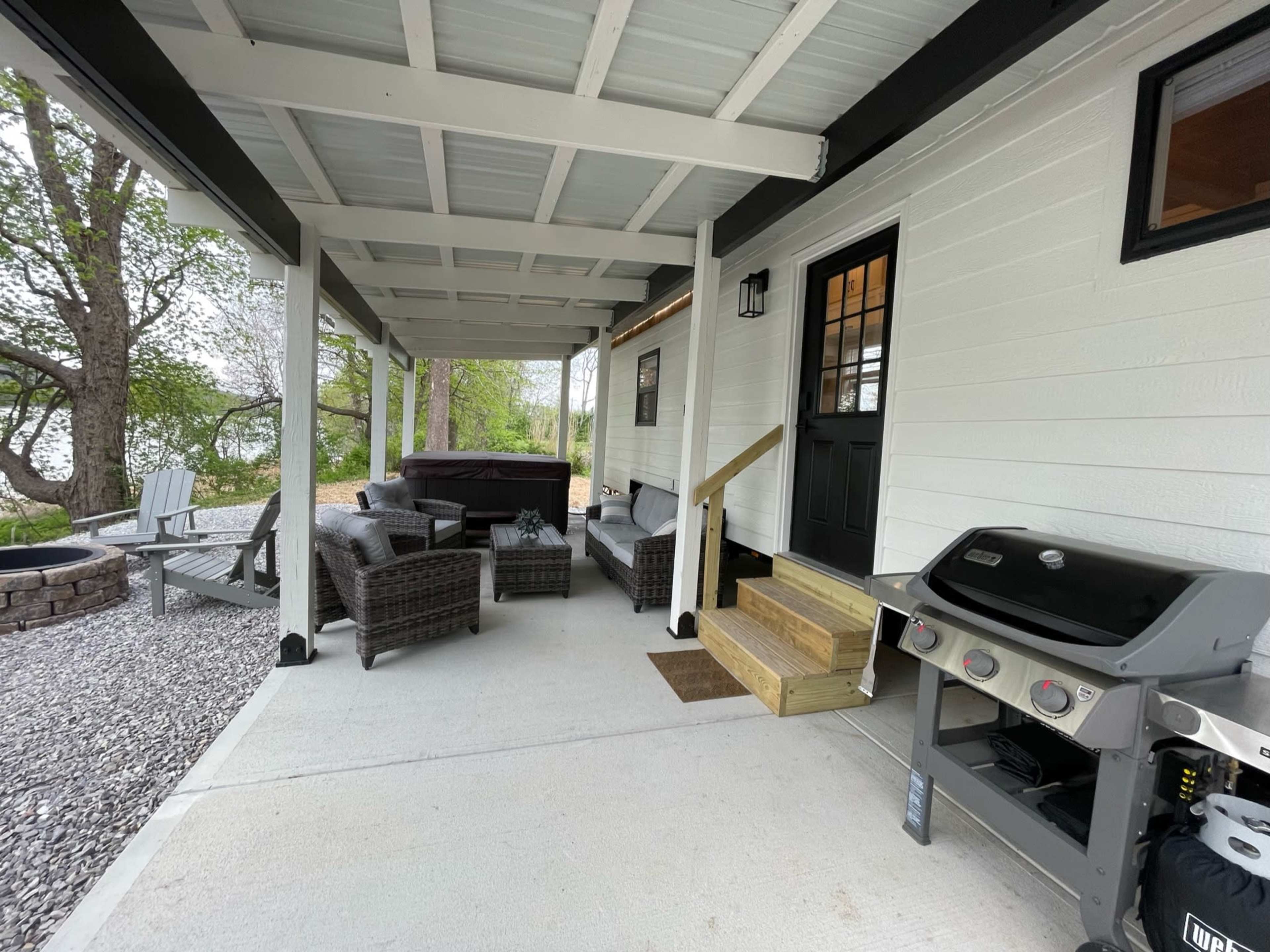 A covered outdoor patio with seating, a barbecue grill, and steps leading to the entrance of a house.