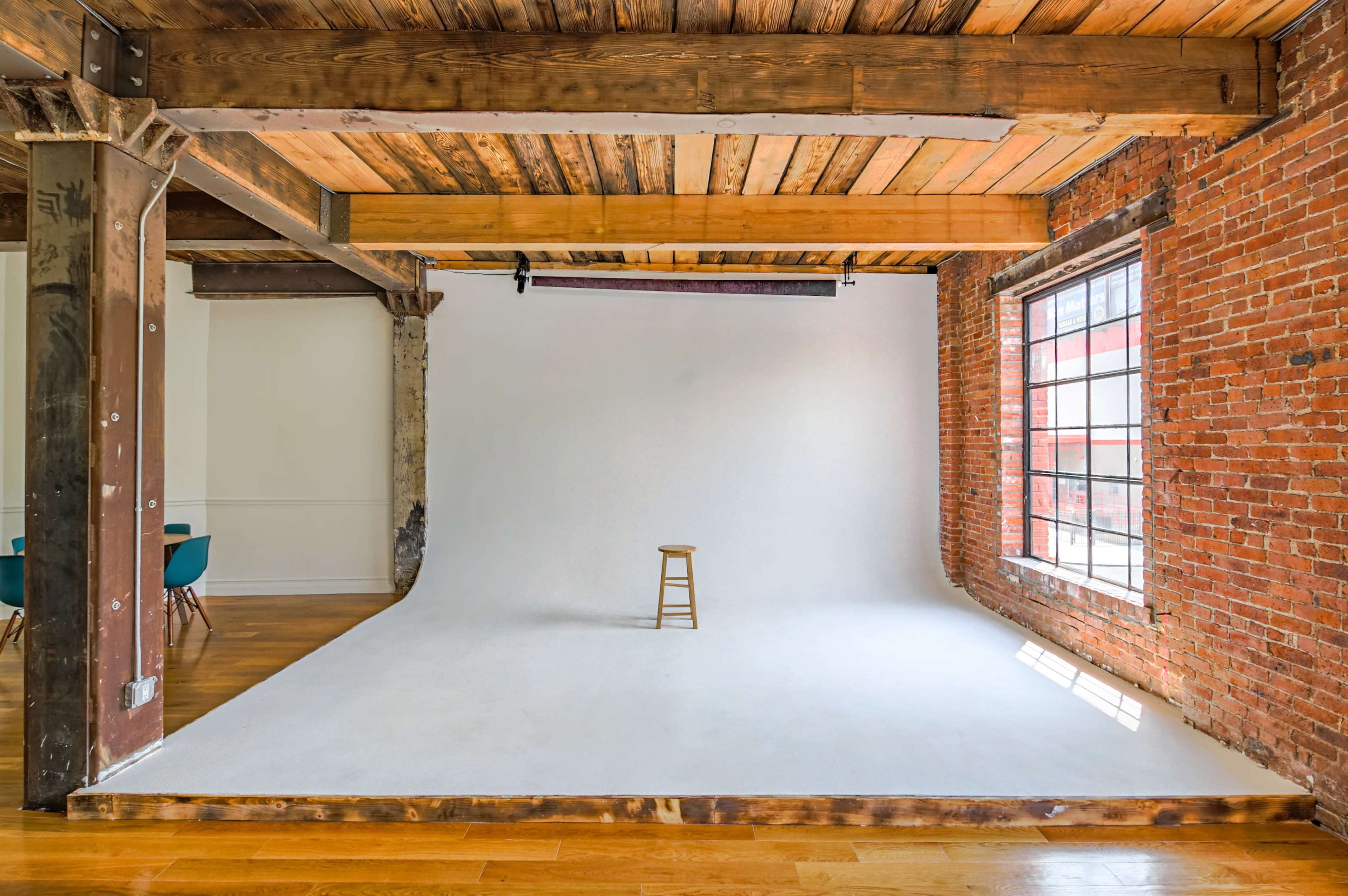 A simple photography studio features a wooden stool on a white backdrop with exposed brick walls and large windows.