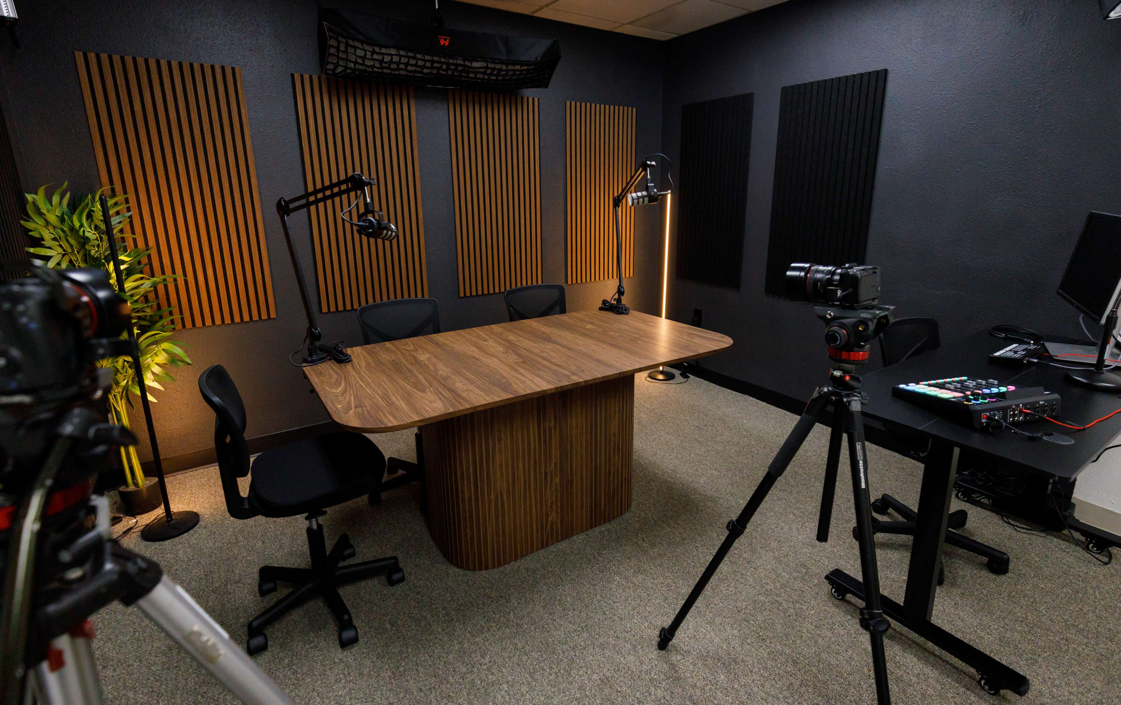 The image shows a studio setup with a large wooden table, two chairs, sound-absorbing panels on the walls, and cameras positioned for recording.