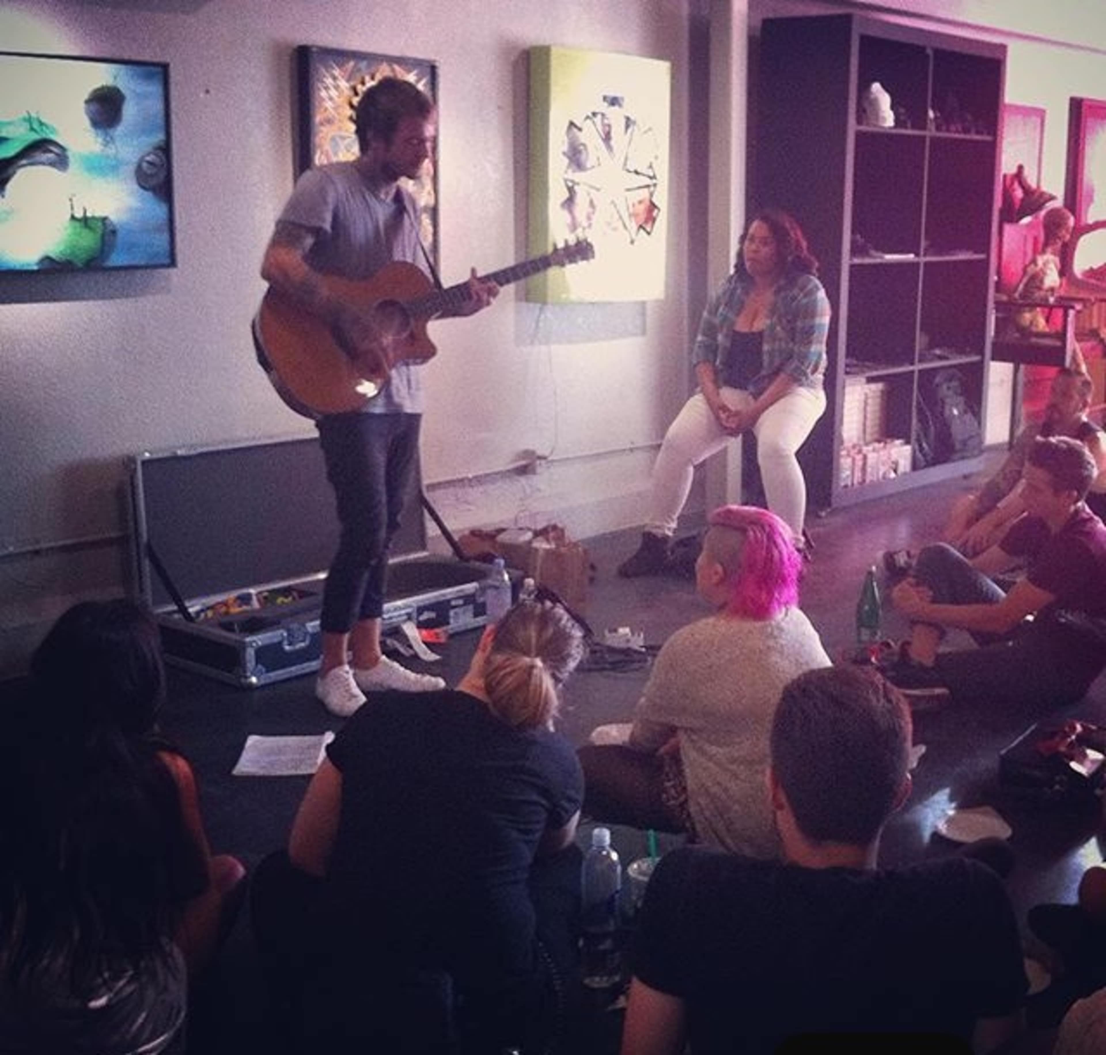 A musician performs with a guitar in front of an audience seated on the floor in an art gallery.