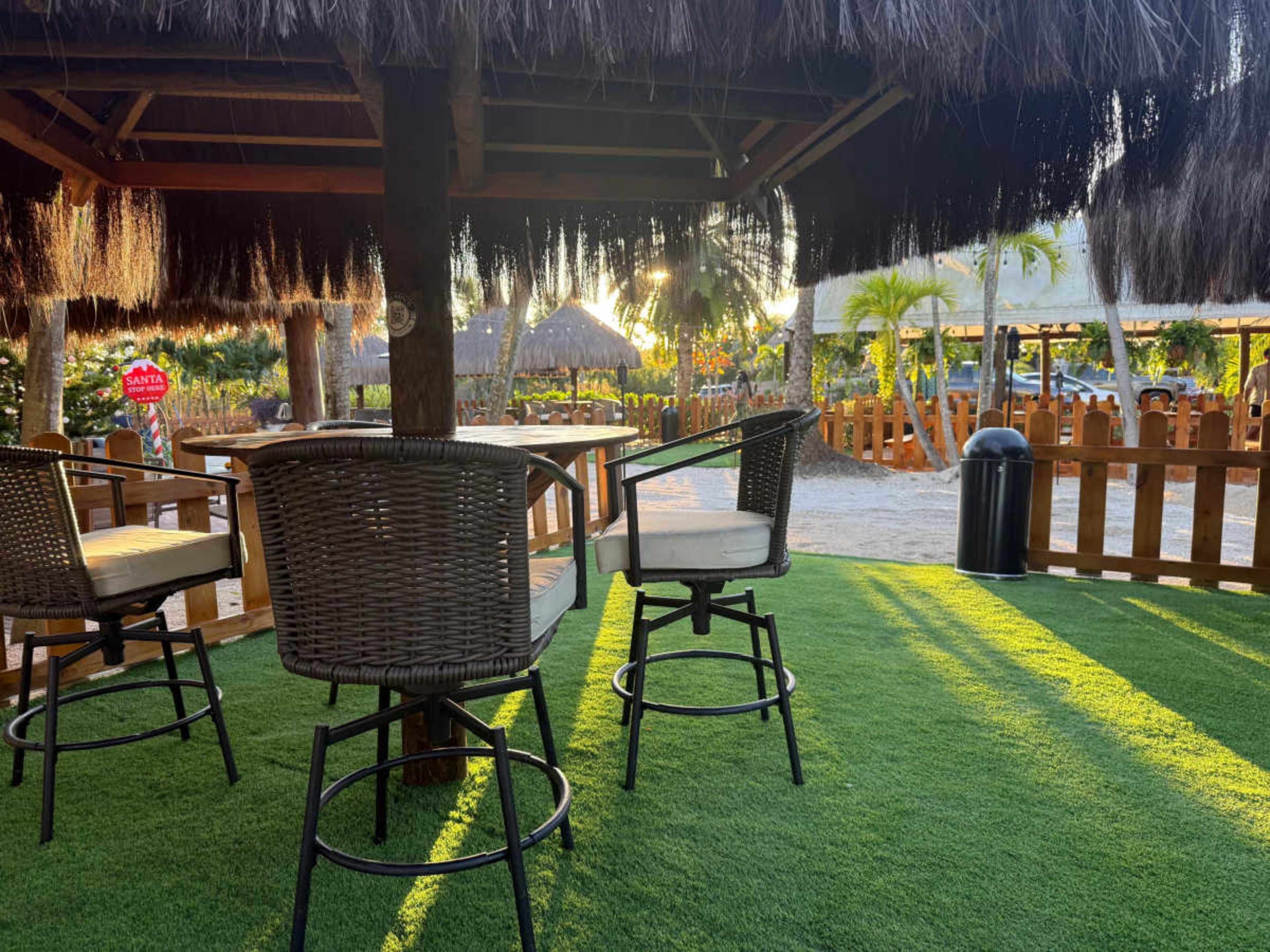 The image shows a shaded outdoor seating area with bar stools, surrounded by greenery and wooden fencing.