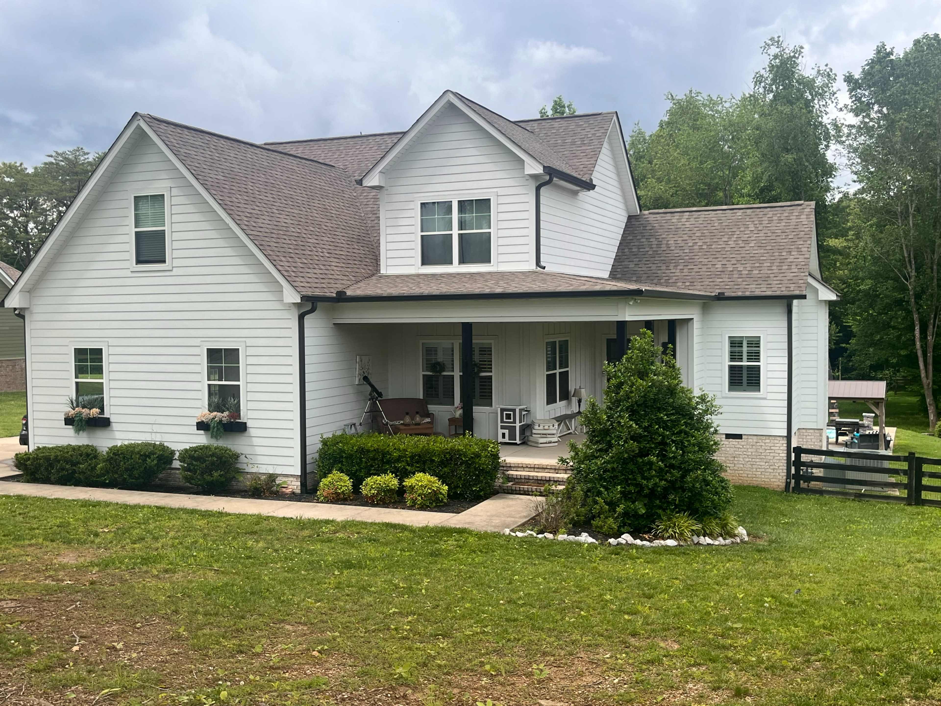 A two-story white house with a front porch, surrounded by green grass and landscaping, features a sloped roof and decorative shutters.