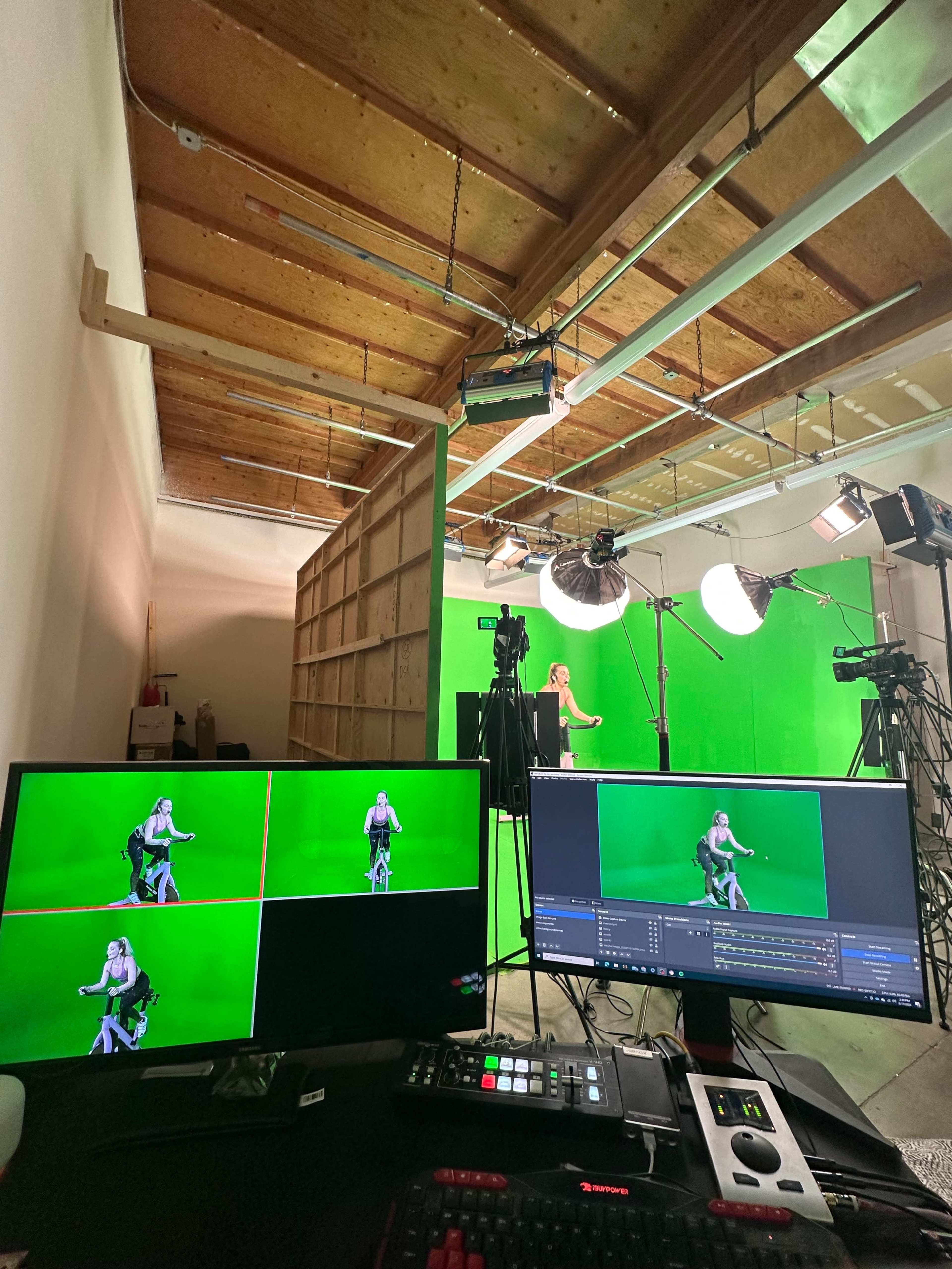 A green screen studio setup with multiple cameras and monitors displaying a person on a stationary bike in front of a green backdrop.