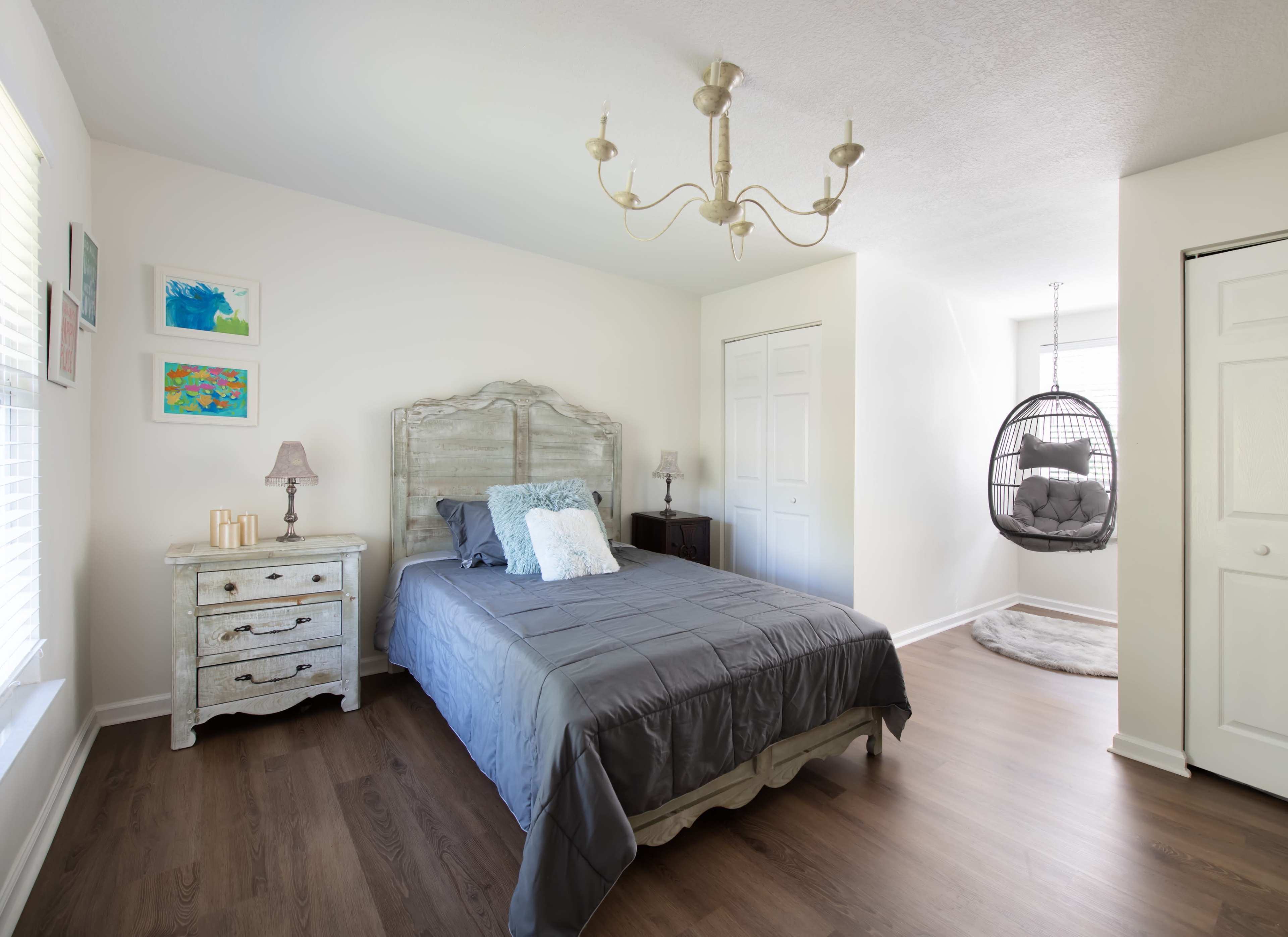 A well-lit bedroom featuring a bed with a gray coverlet, a chandelier, and a hanging chair near a closet.