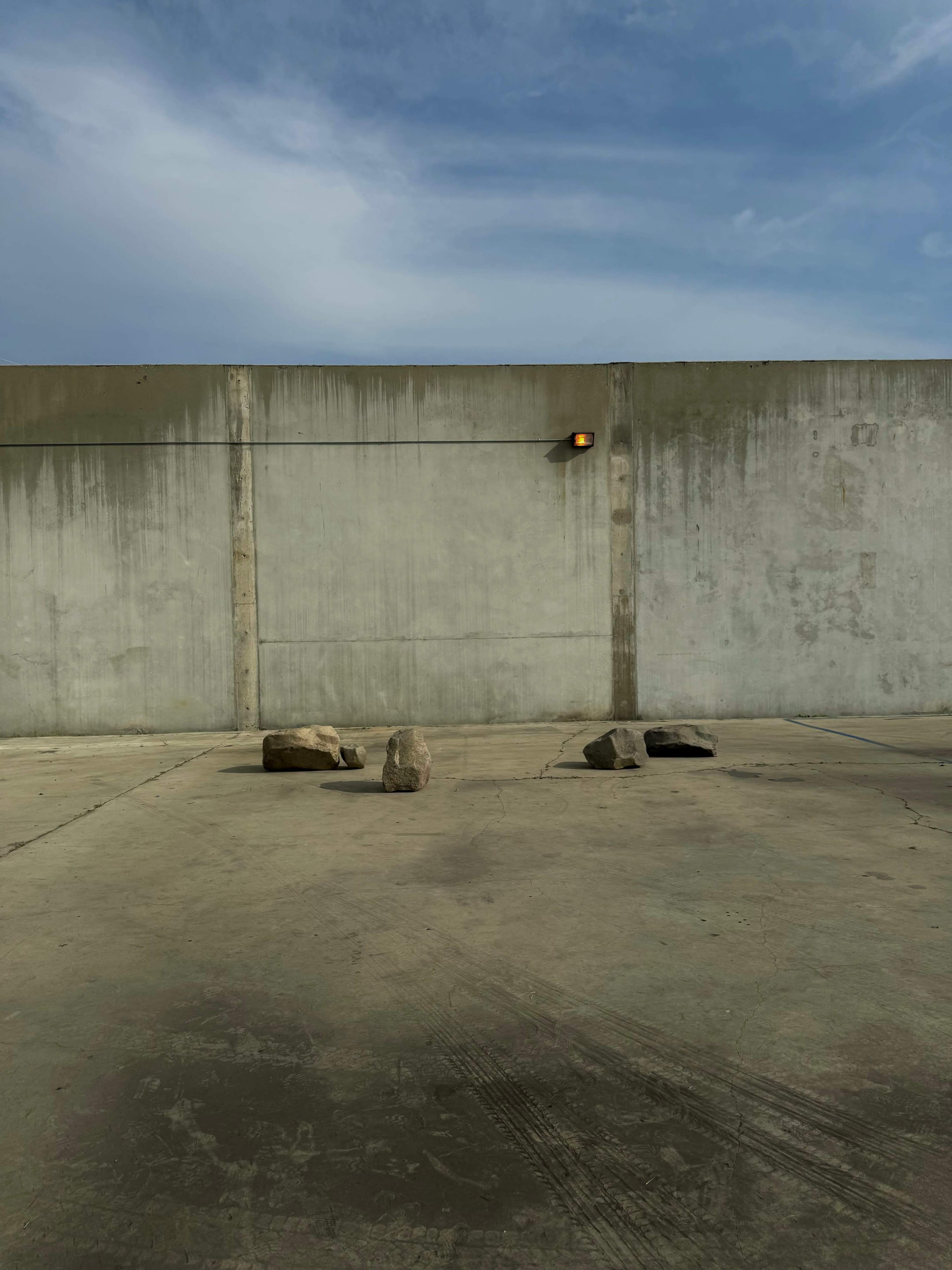 A concrete surface with four rocks arranged in a row in front of a plain wall under a blue sky.