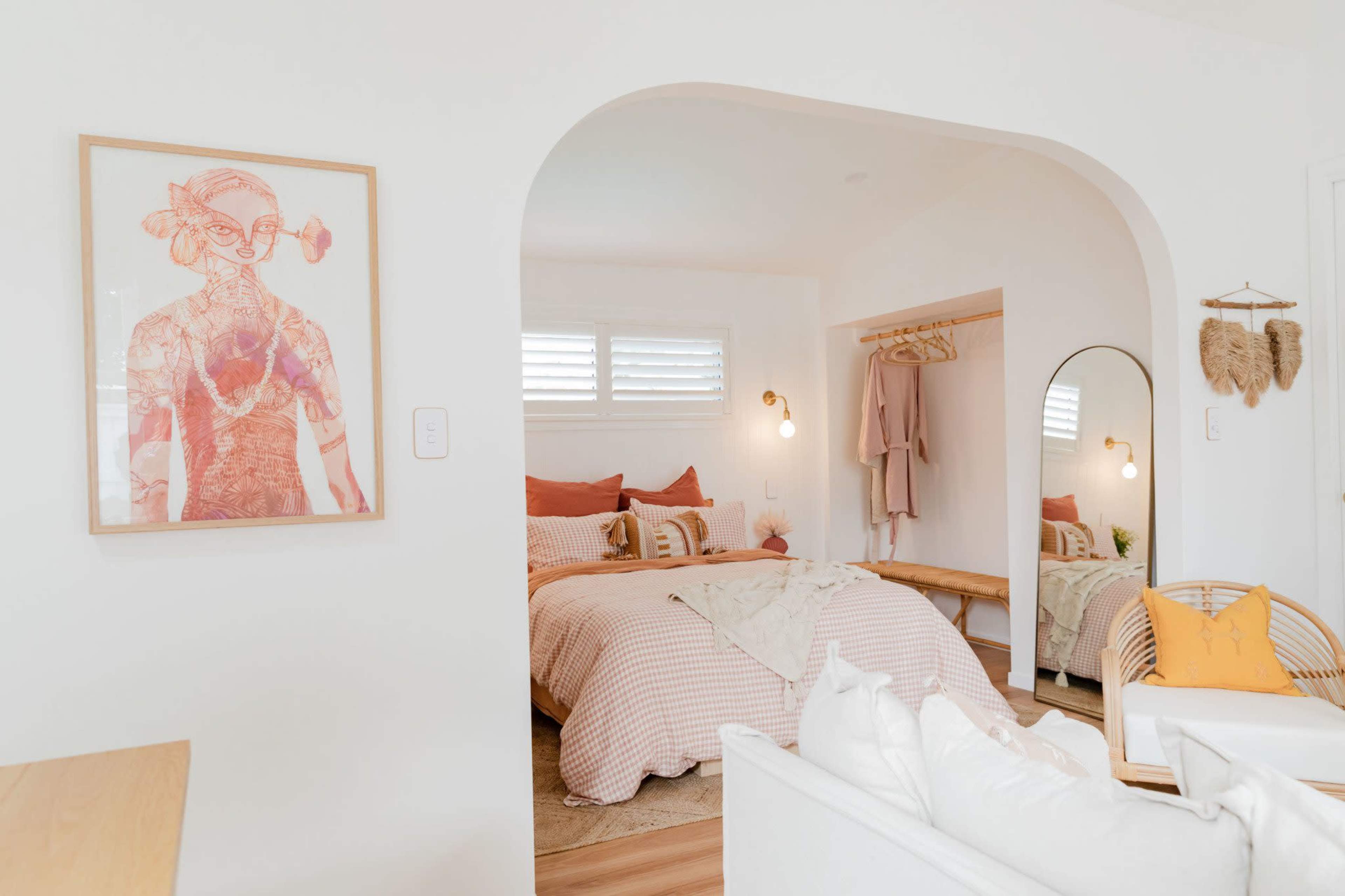 The image features a light-filled bedroom with a bed dressed in patterned linens, an archway leading to a sitting area, and a large mirror along with a coat rack mounted on the wall.