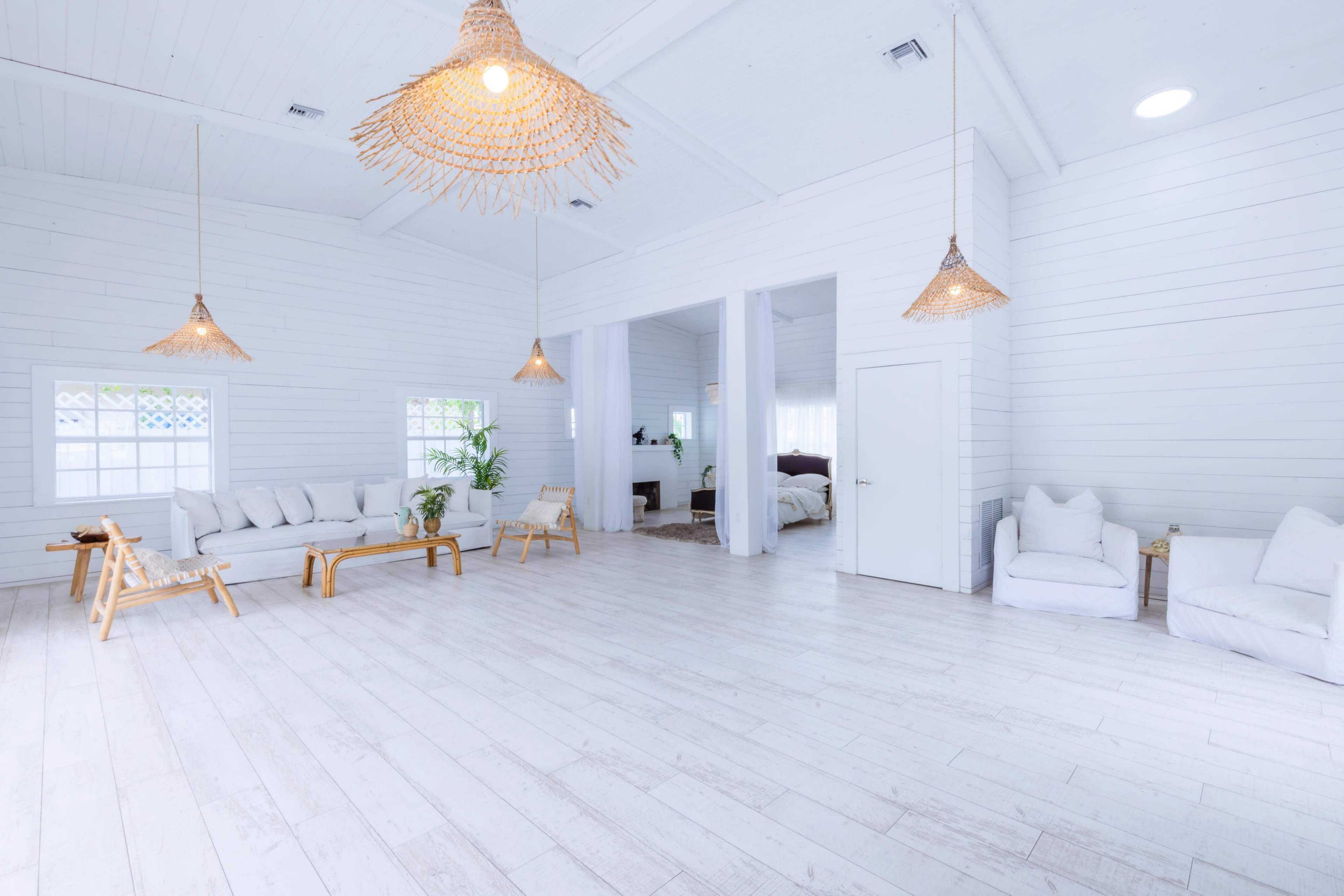 A bright, open room features white walls and wooden flooring, furnished with a sofa, chairs, and hanging woven light fixtures.