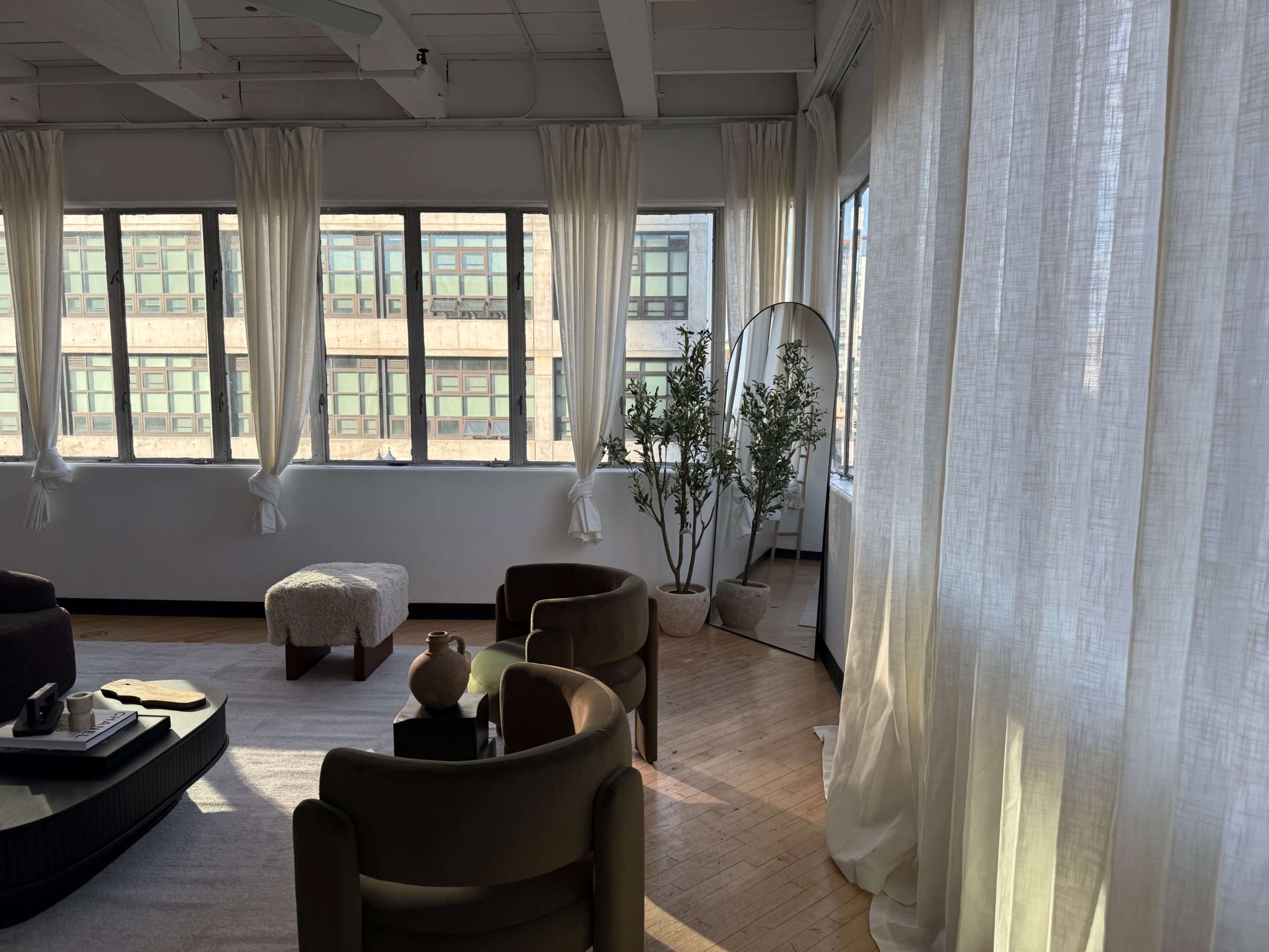 A well-lit room features large windows draped with sheer curtains, two chairs, a small ottoman, and a potted plant in the corner.