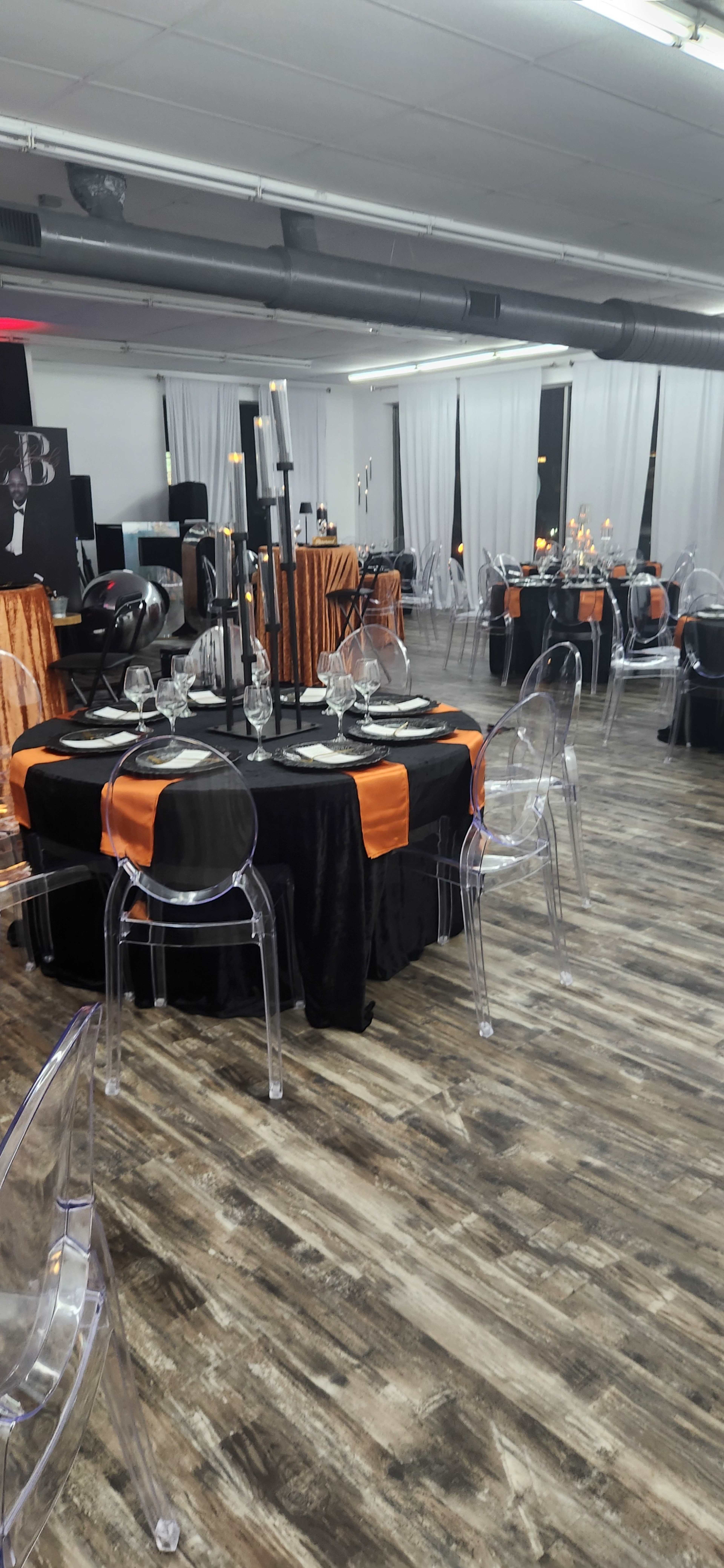 The image shows a banquet hall set up with tables draped in black and orange, featuring clear acrylic chairs and decorative centerpieces.