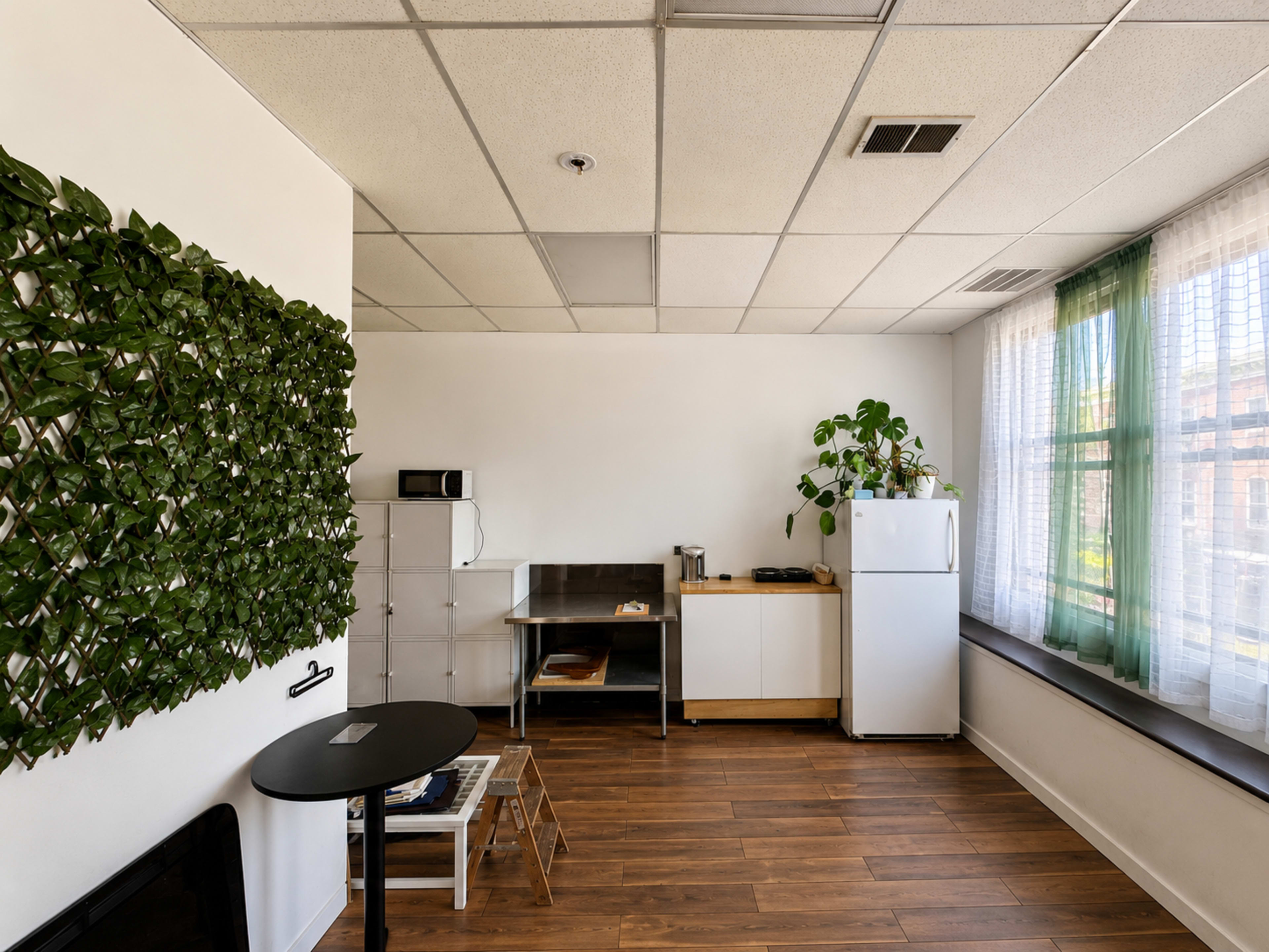 The image shows a modern kitchen area with a wooden floor, a white refrigerator, a small dining table, and a wall adorned with green plants.