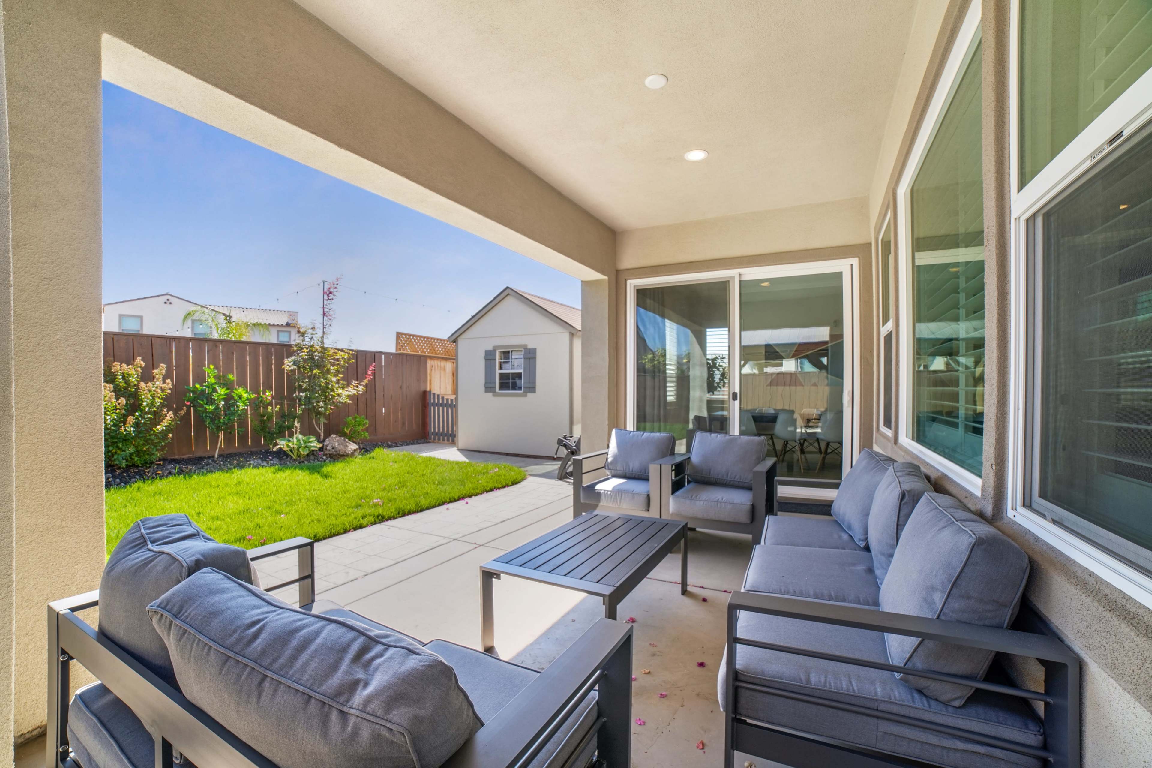 A covered patio features a seating area with cushioned chairs and a coffee table, overlooking a neatly landscaped backyard with a shed.