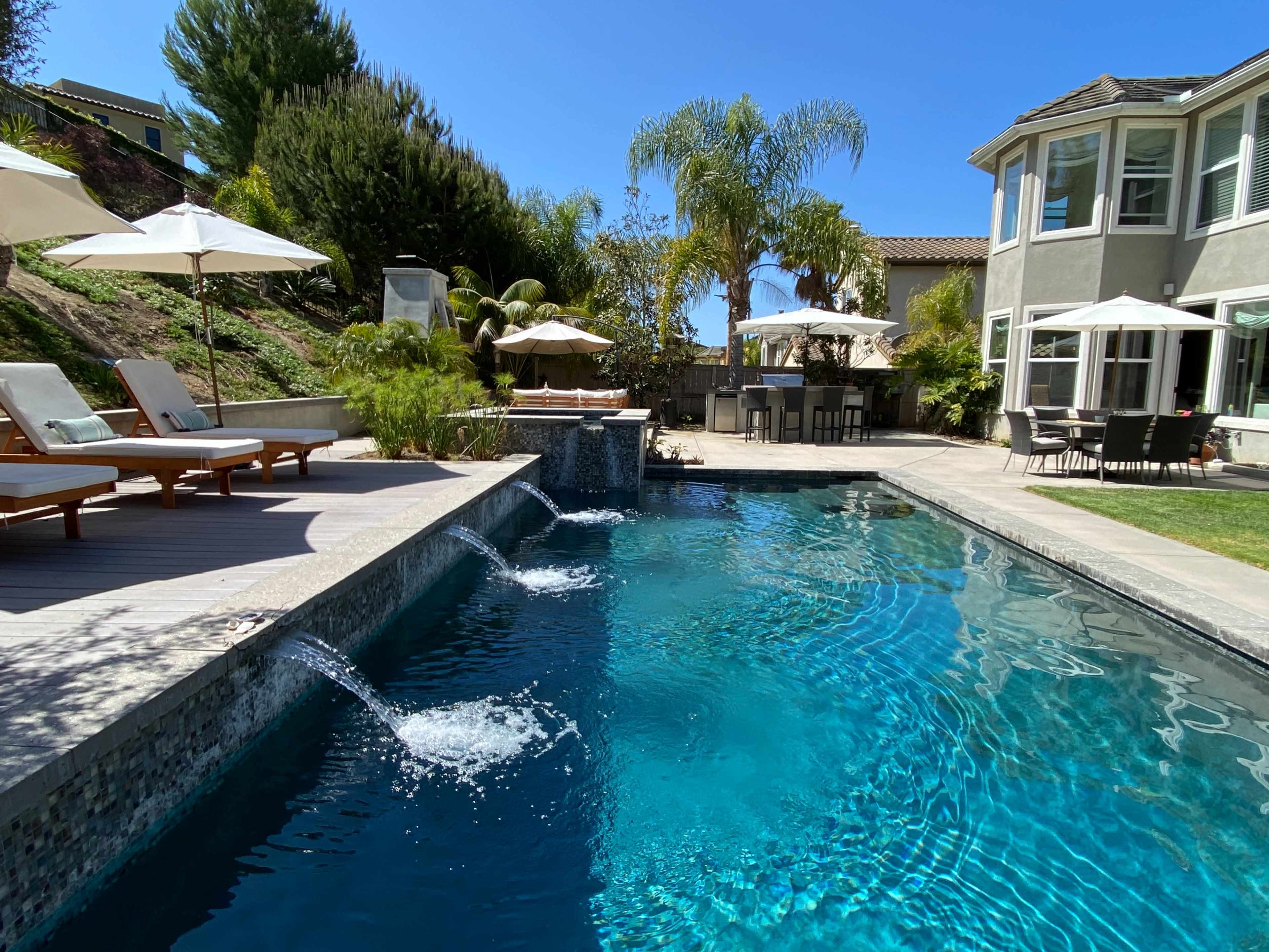 A residential backyard with a swimming pool featuring waterfall fountains, surrounded by lounge chairs, parasols, and a dining area.
