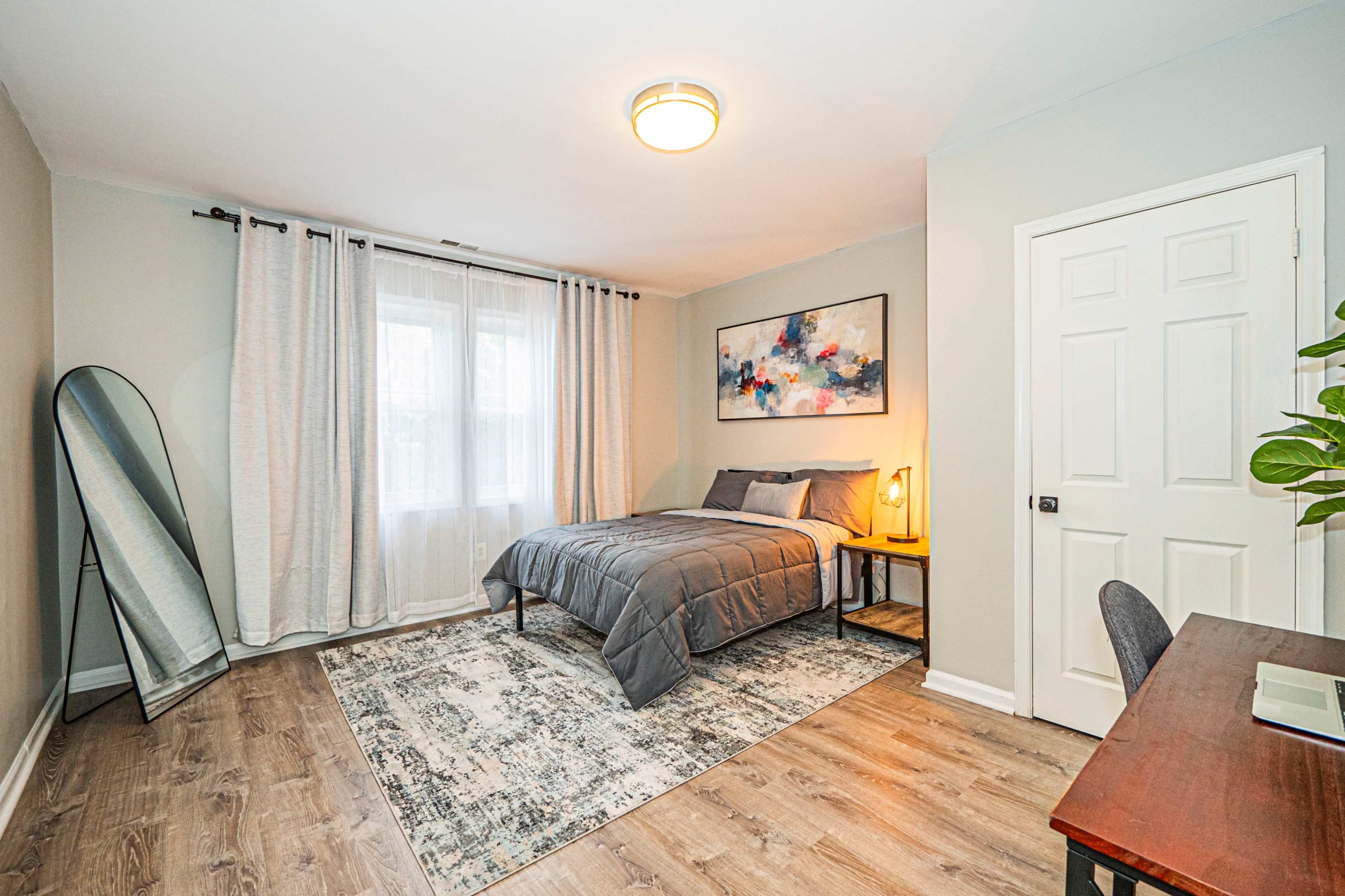 The image shows a simple bedroom with a bed, a desk, a large mirror, and a window dressed with curtains.