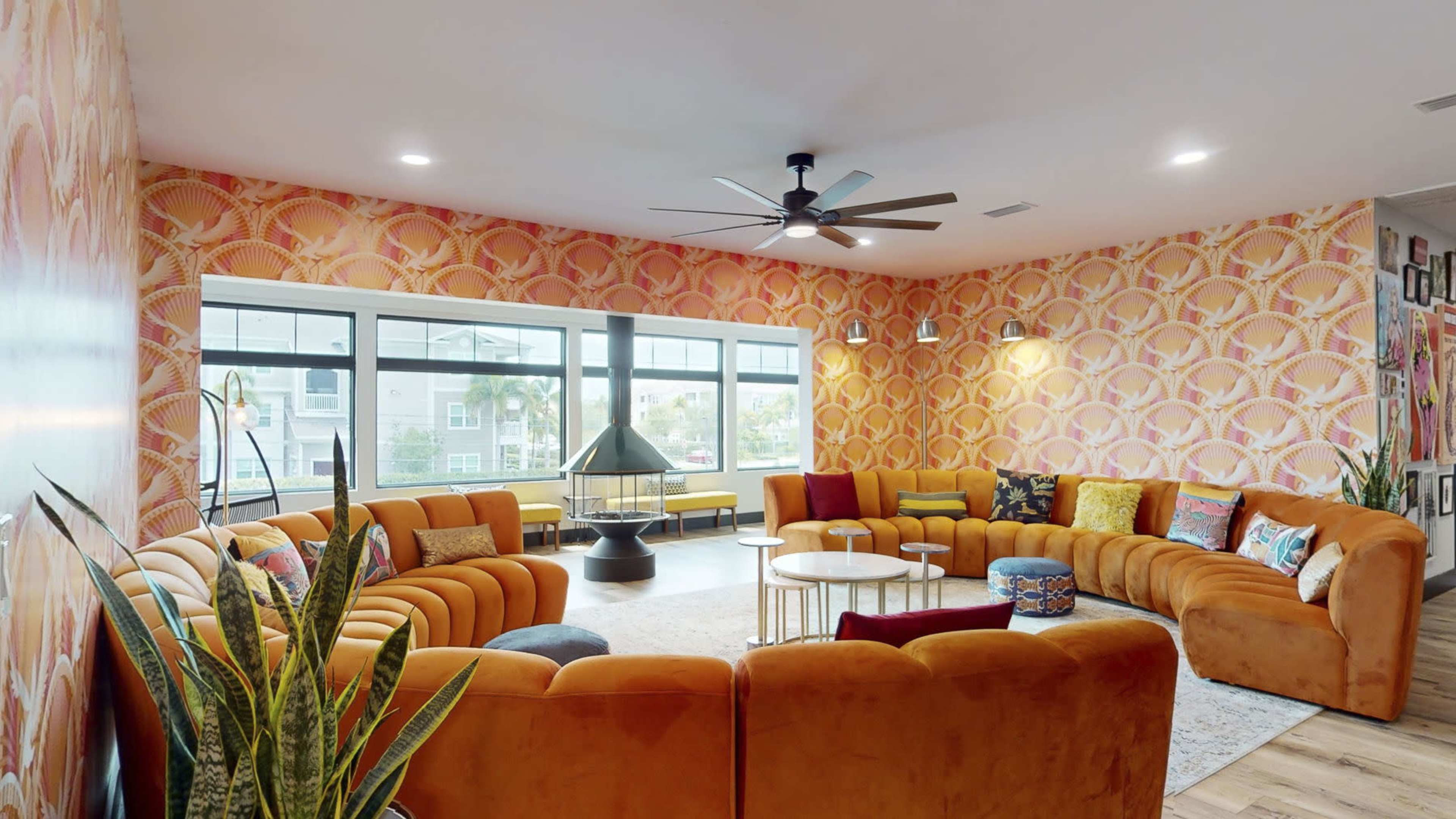 A brightly colored lounge features a circular arrangement of orange sofas around a central coffee table, with large windows letting in natural light.