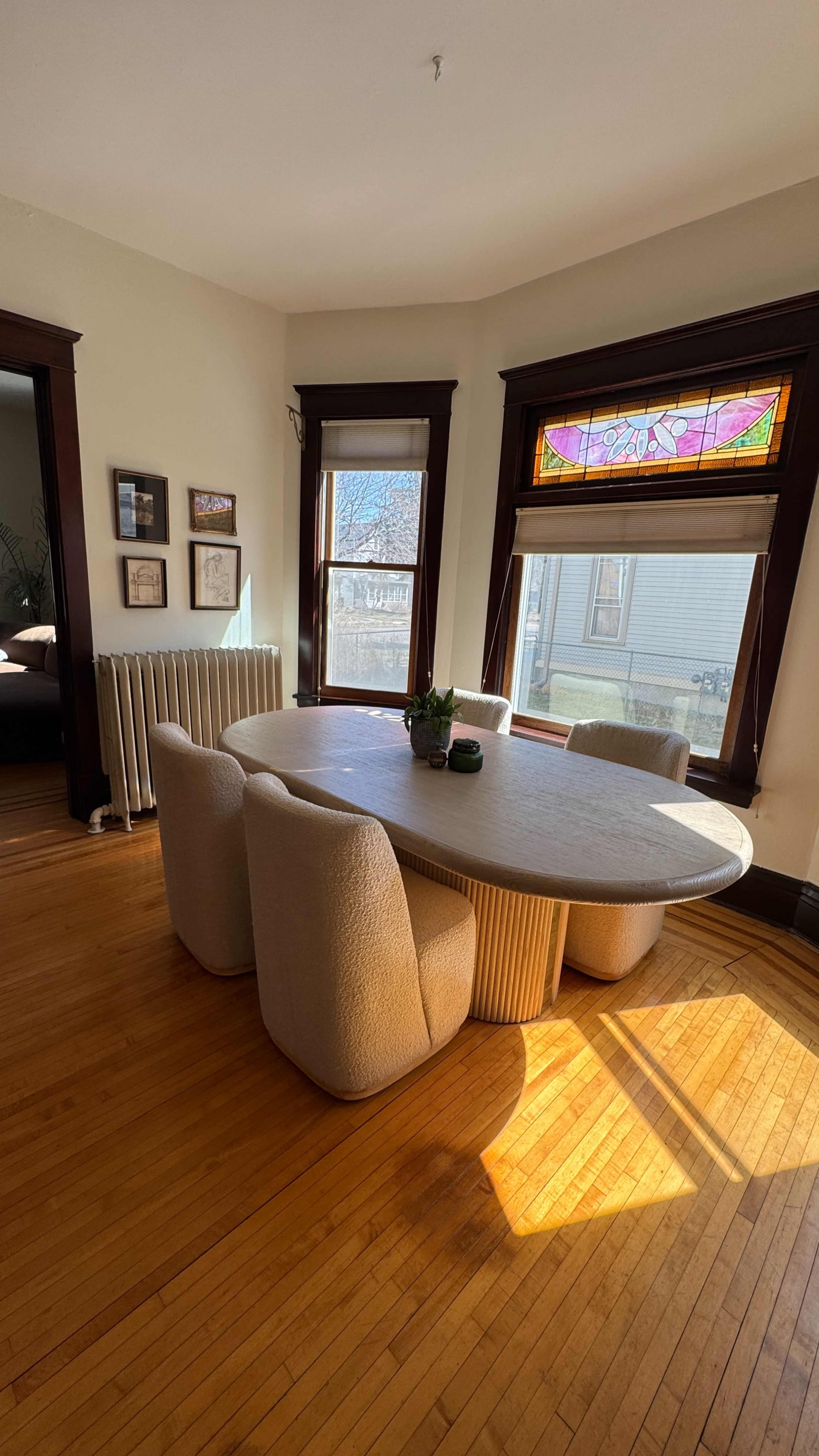 A round dining table surrounded by upholstered chairs is illuminated by sunlight streaming through stained glass windows in a cozy room.