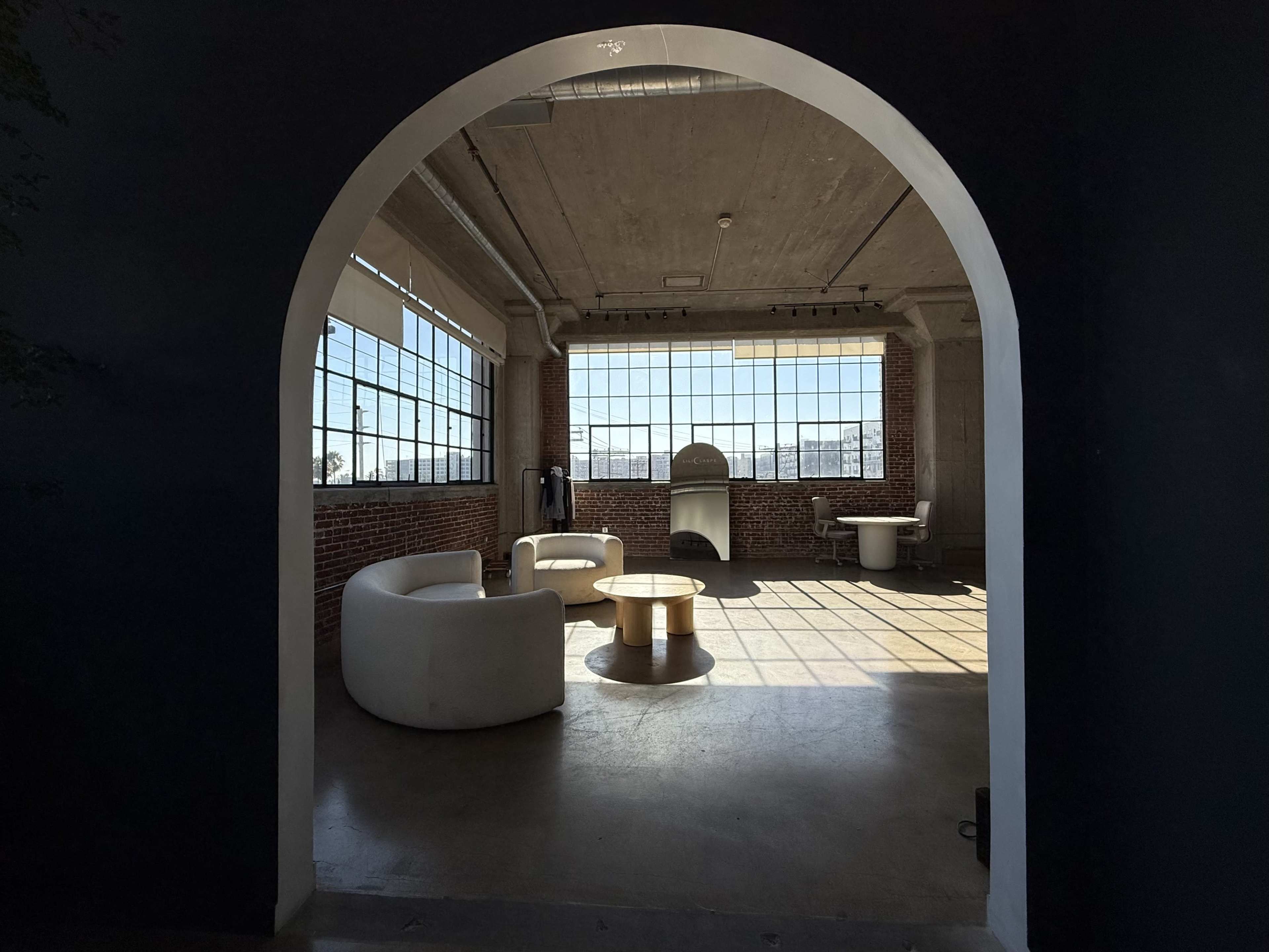 The image shows a spacious, industrial-style room with large windows, featuring a circular table and two white chairs.
