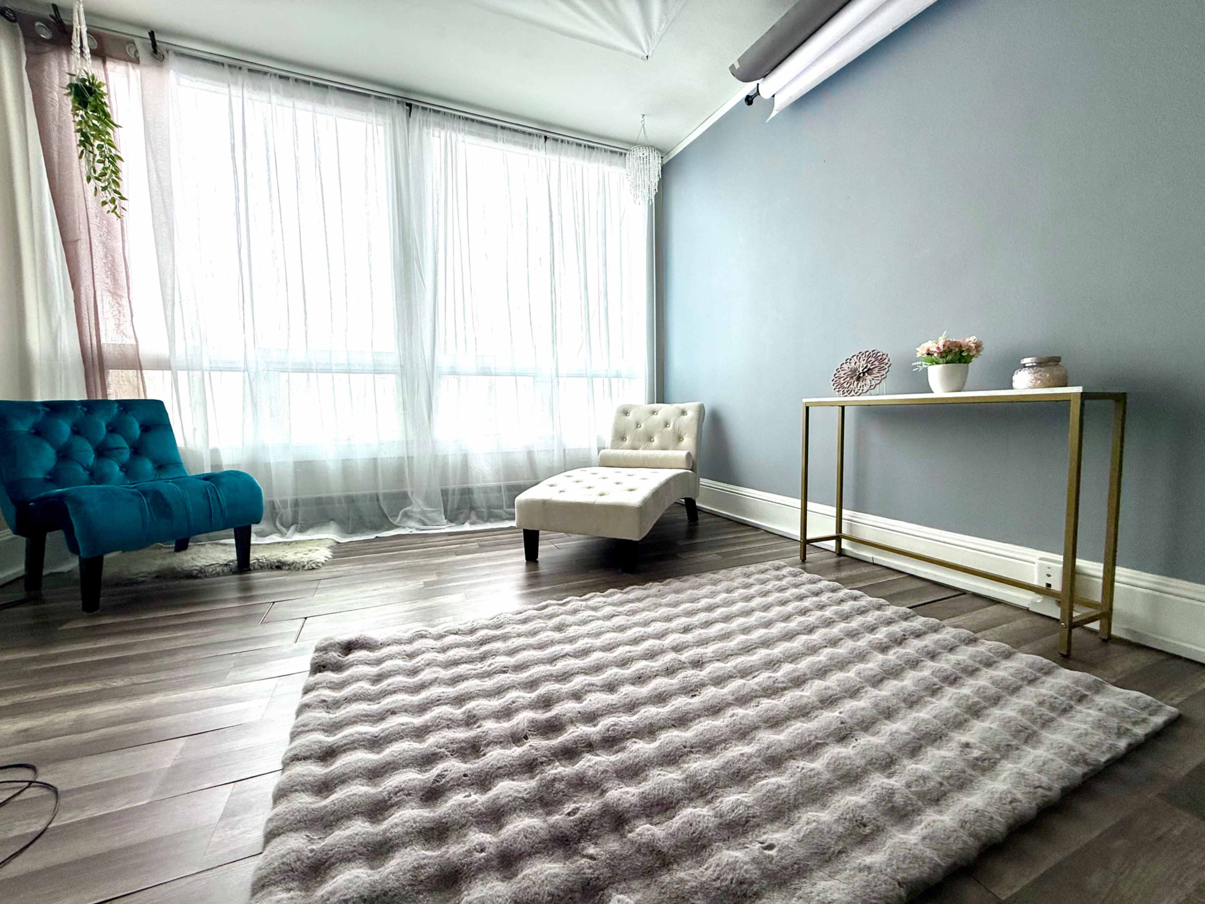 The image features a minimalist room with large windows, light-colored curtains, a blue armchair, a white upholstered chair, and a gold side table adorned with decorative items.