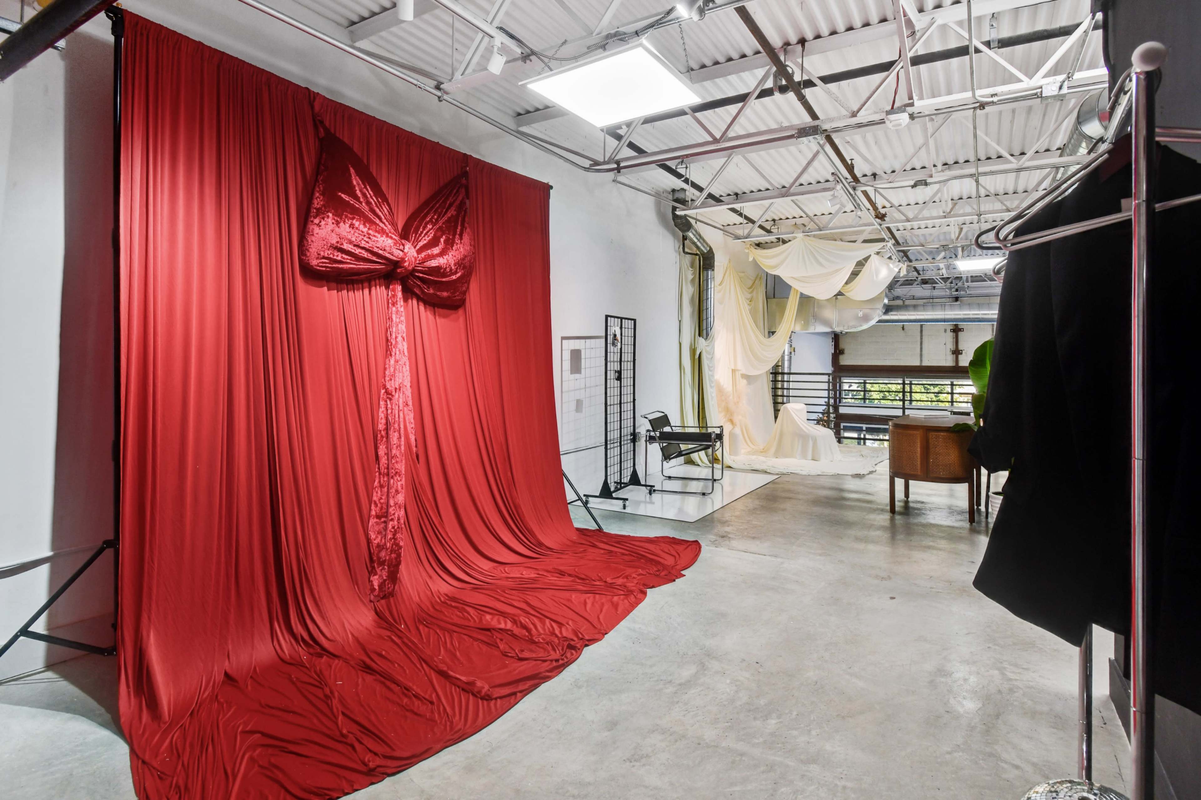 The image shows a spacious studio with a large red fabric backdrop styled as a bow, complemented by draped white fabric in the background and various furniture pieces arranged throughout the space.