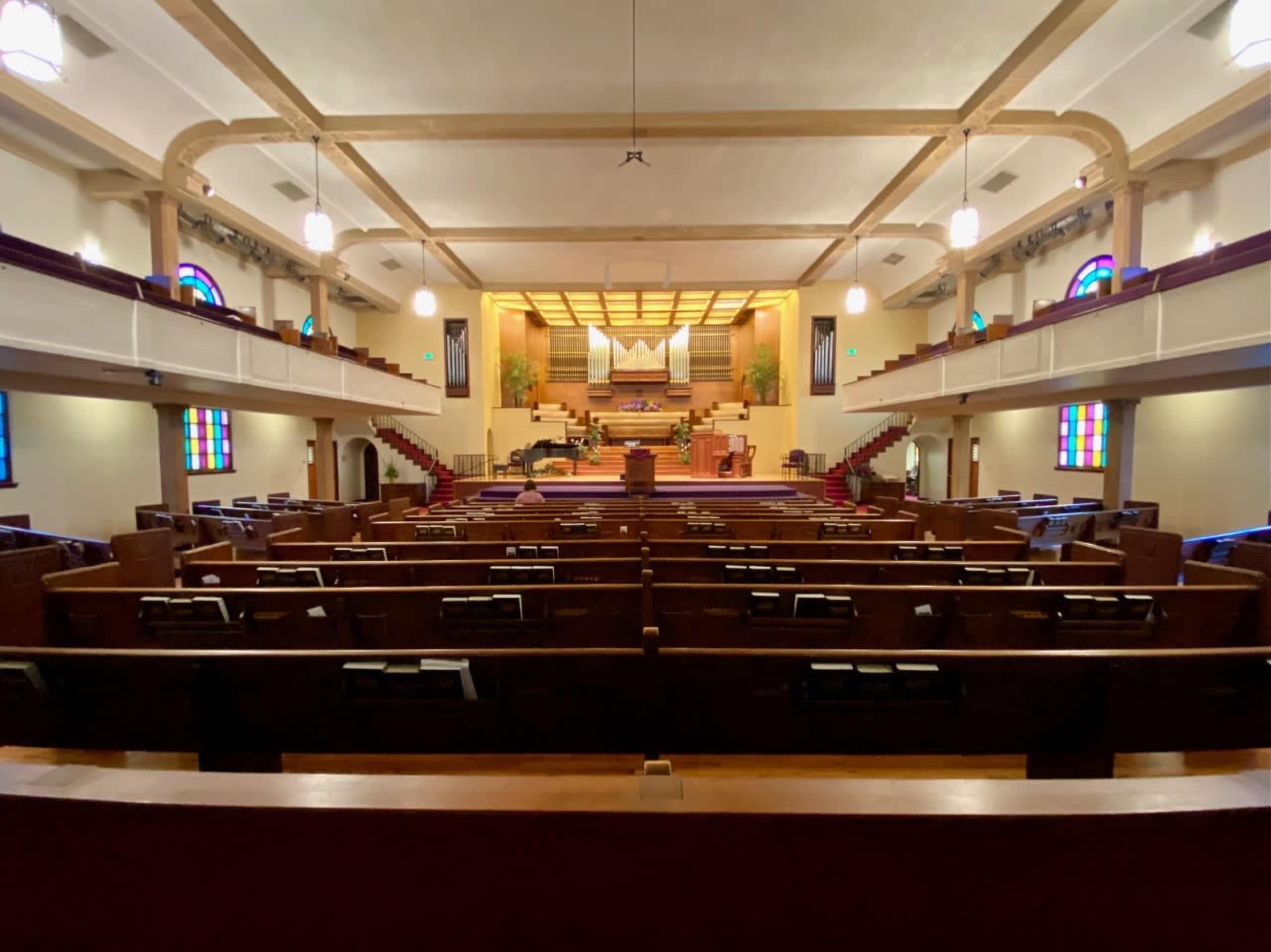The interior of a spacious church with wooden pews and a stage at the front, featuring stained glass windows.