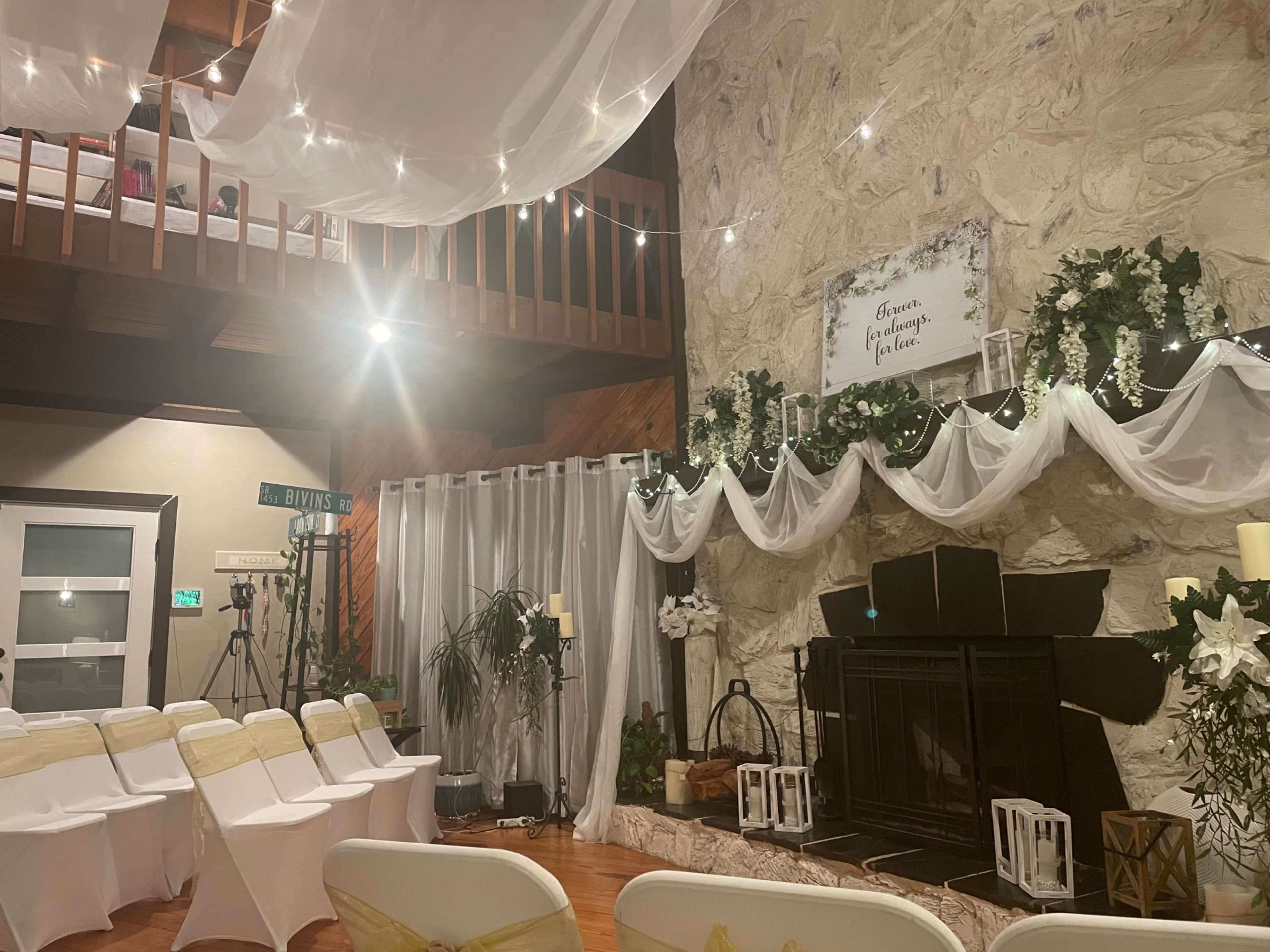 The interior of a venue features a stone wall with a fireplace, adorned with draped fabric and fairy lights, while rows of white chairs with yellow sashes are set up for an event.