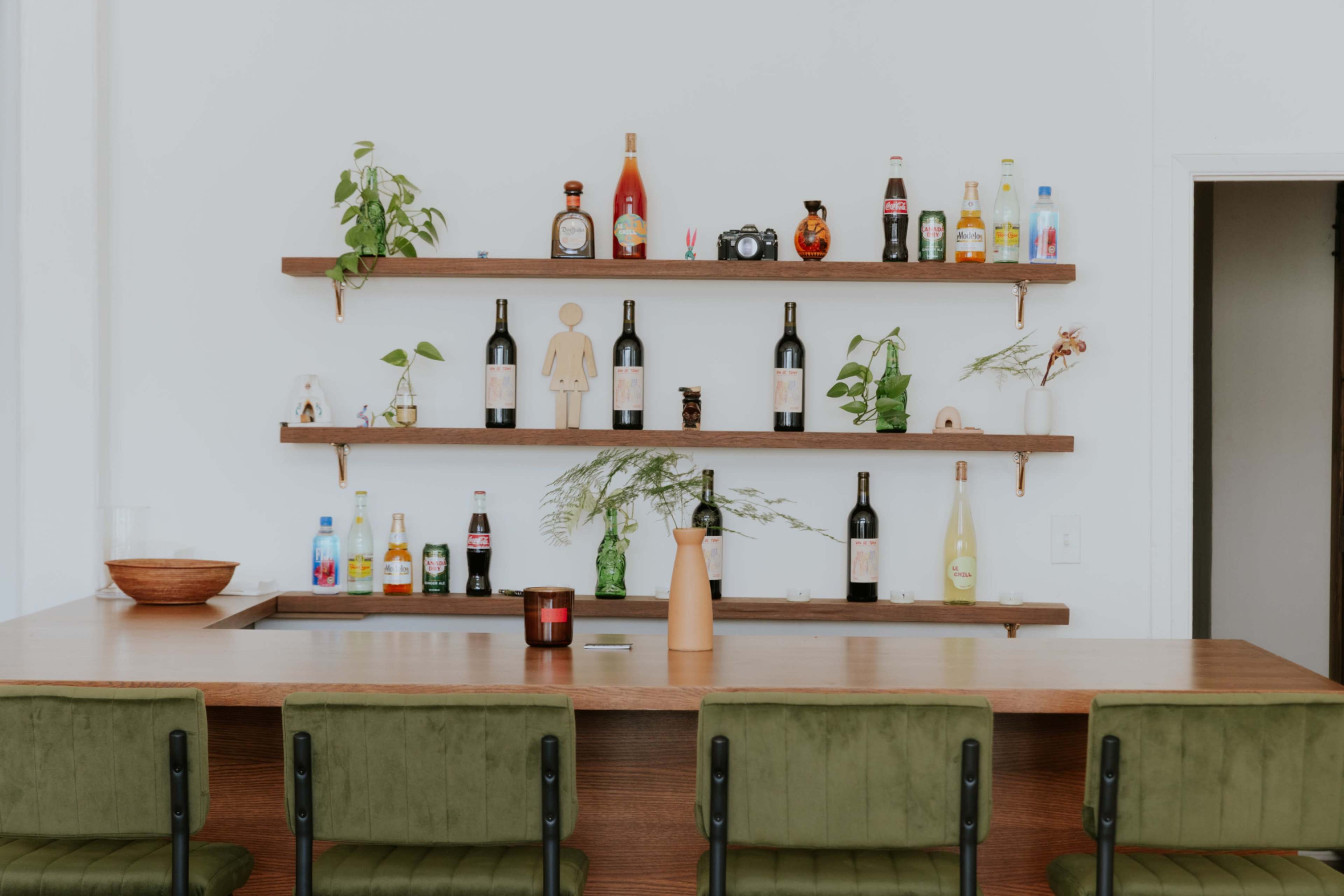 A wooden bar with green chairs displays a variety of bottles and decorative items on shelves above.