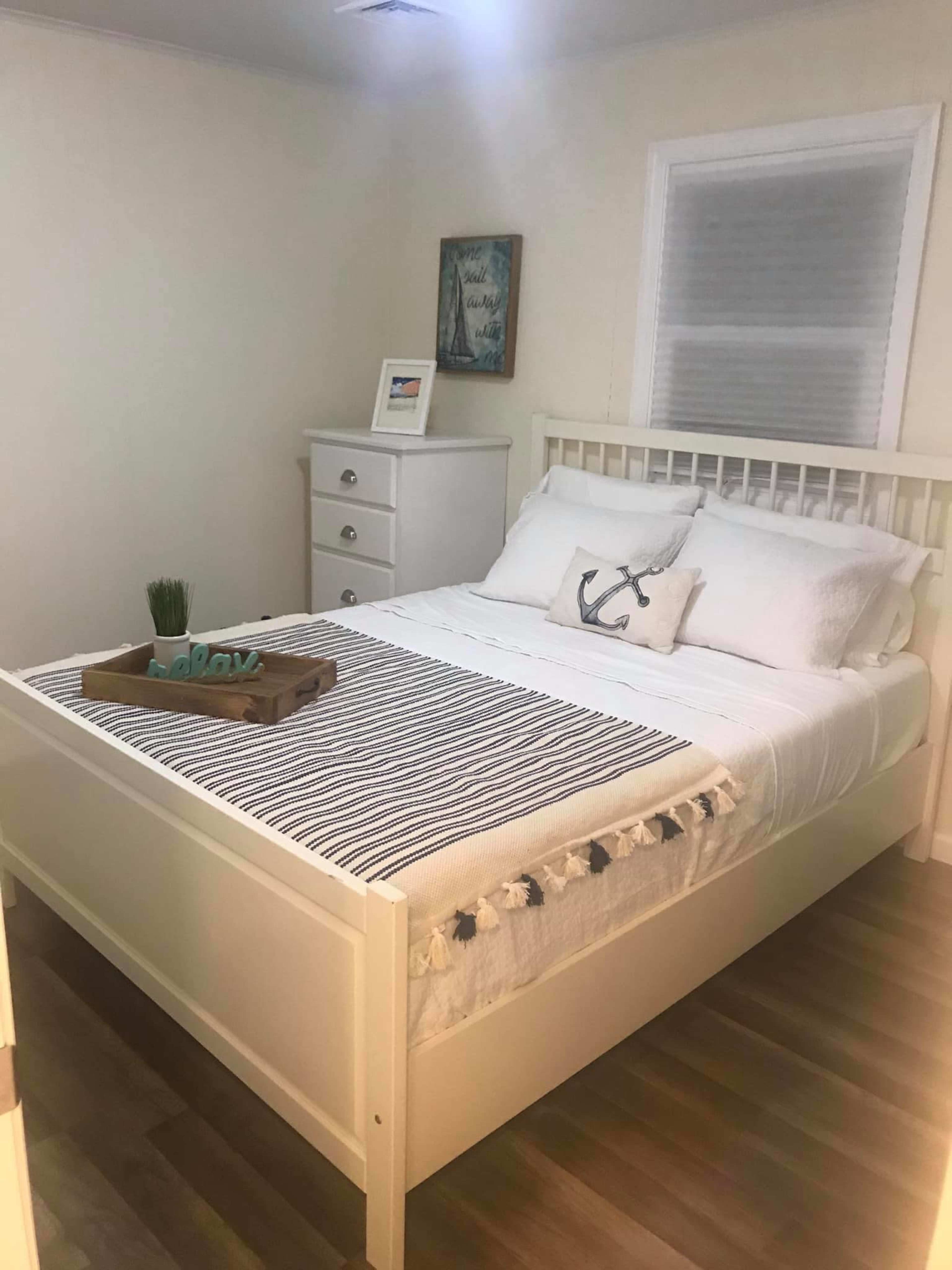 The room features a white bed with a striped throw blanket, accompanied by a wooden tray on the bed, and a light-colored dresser beside it.