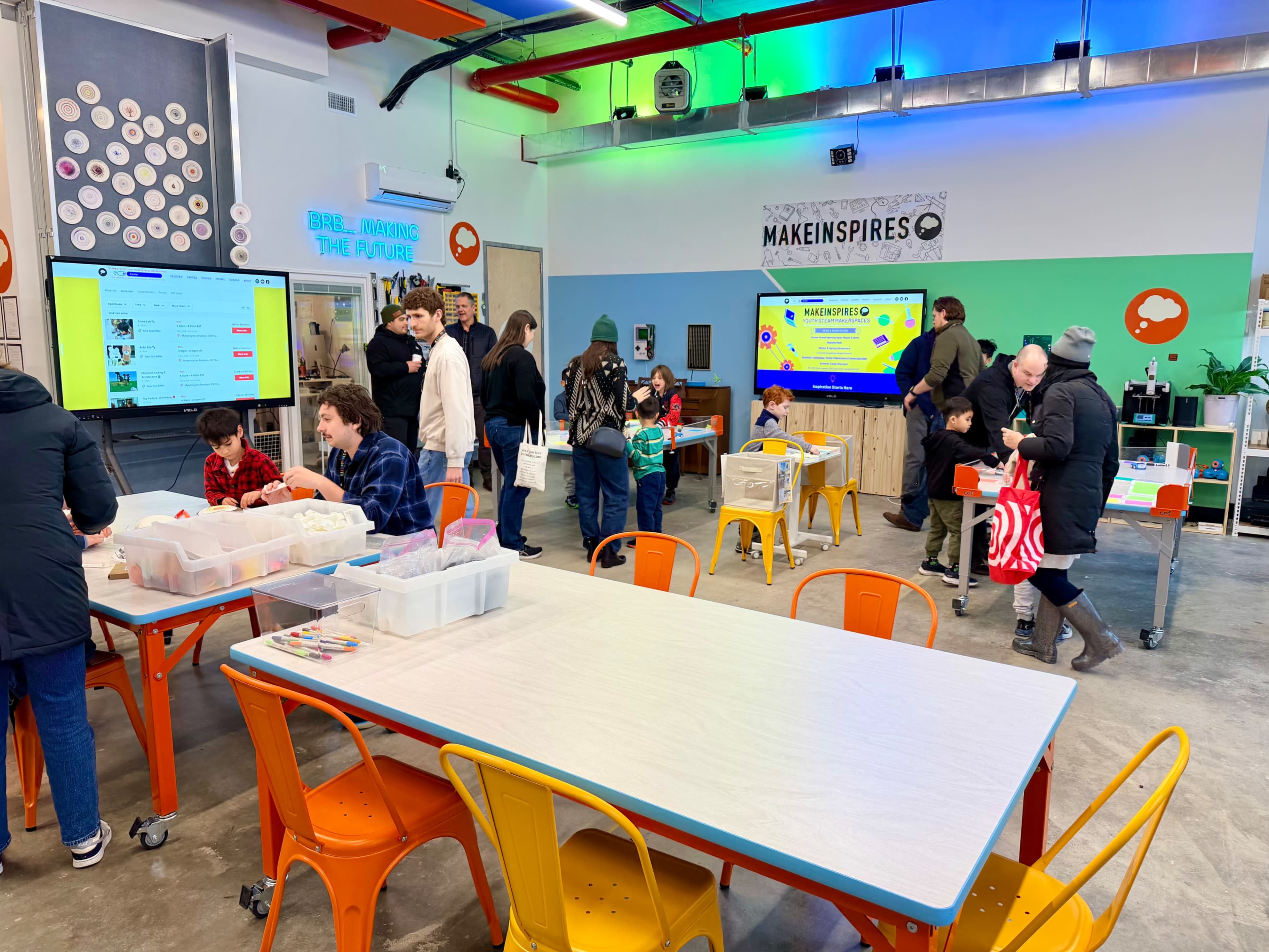 A bright, modern interactive workspace features children and adults engaging in activities at colorful tables, with screens displaying information on the walls.