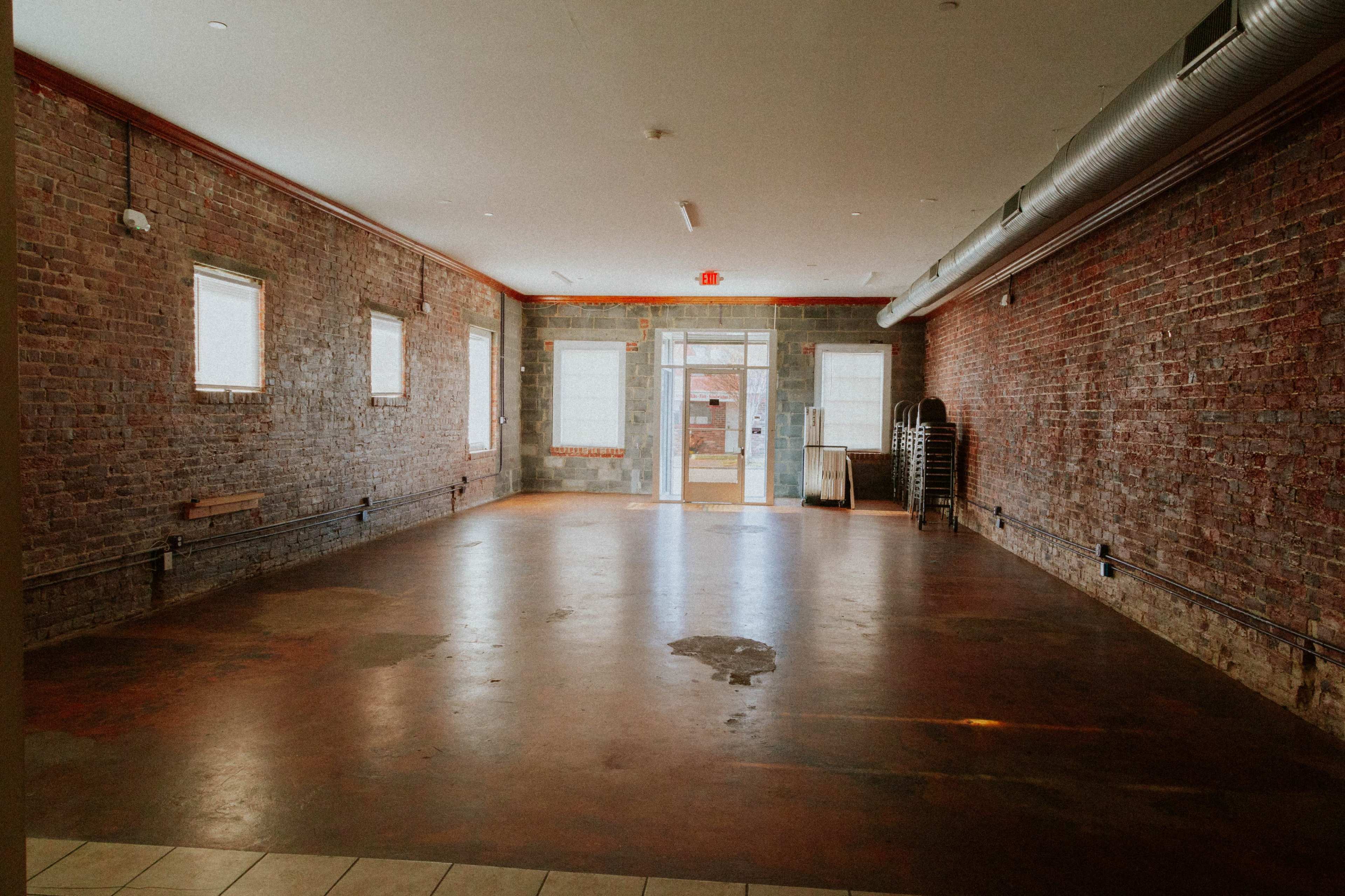 The image shows an empty room with brick walls and a concrete floor, featuring windows and a doorway leading outside.